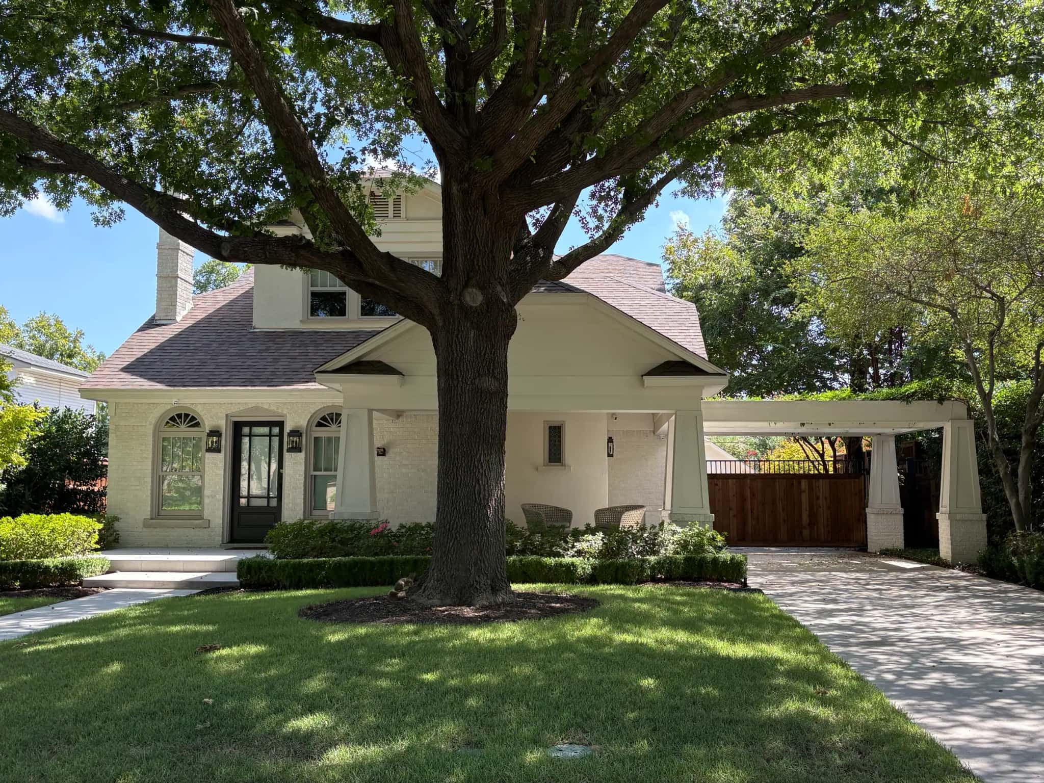 A white-painted brick bungalow under a large spreading tree: a deep front porch on tapered columns, a black front door with arched windows either side, and a matching porte-cochere over the drive.