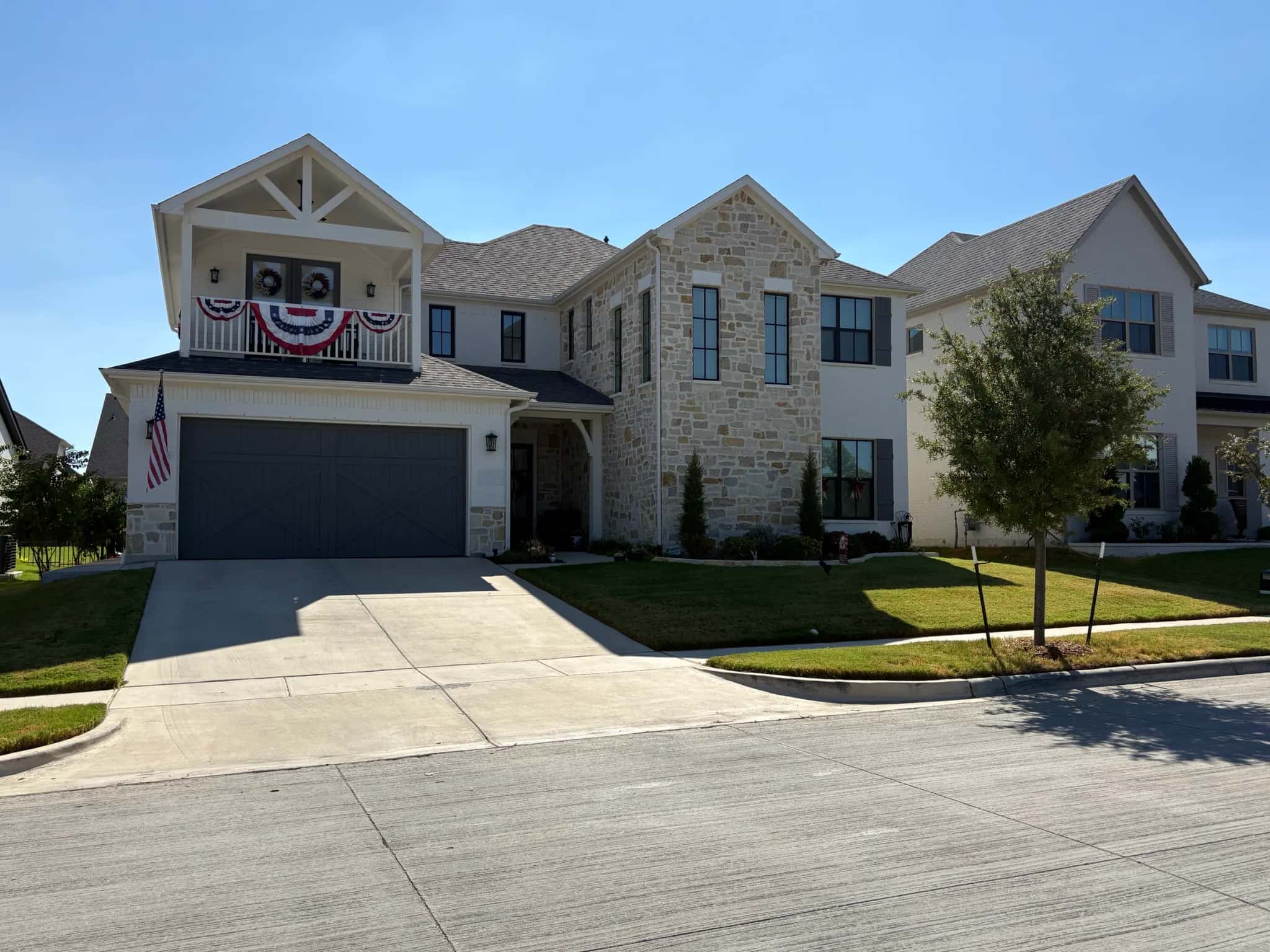 A two-story house of cream stone and white brick with a covered balcony over a gray carriage-style garage door, seen from across the street.