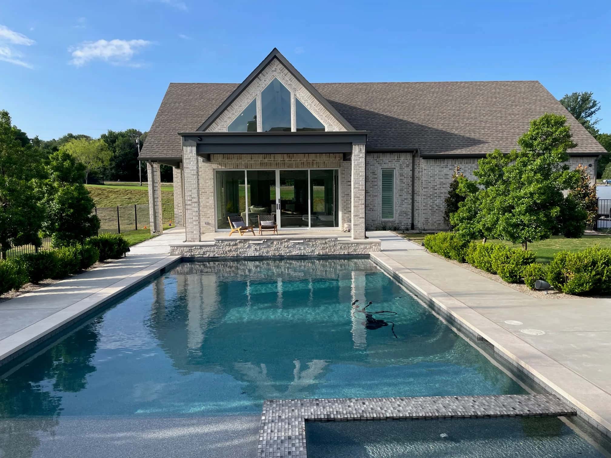 A rectangular pool with a raised spa and a tile-edged tanning ledge, looking back at a blond-brick house with a tall glass gable over a covered patio and sliding doors.