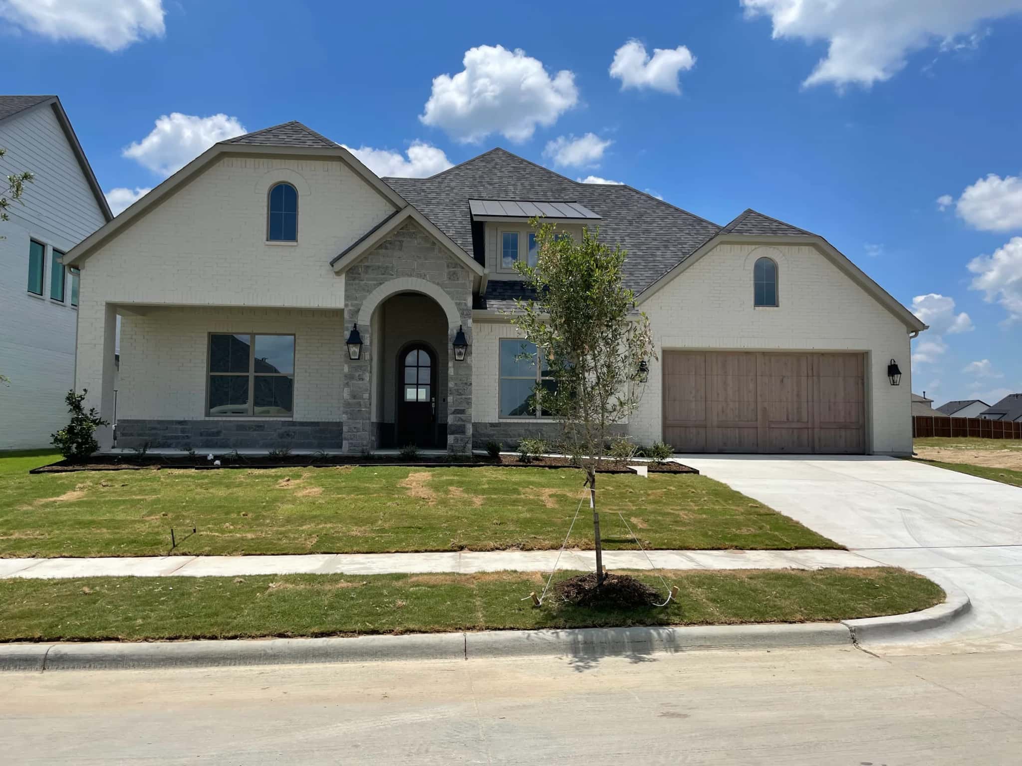 A single-story white brick house with a stone arched entry, arched windows, a standing-seam accent roof and a wood two-car garage door, freshly sodded.