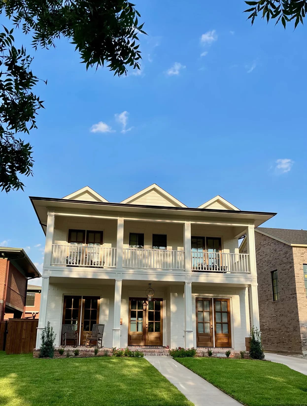 A two-story white-painted brick house with a full-width gallery porch on each floor, wood French doors and a gas lantern over the entry, seen from the front lawn in low sun.