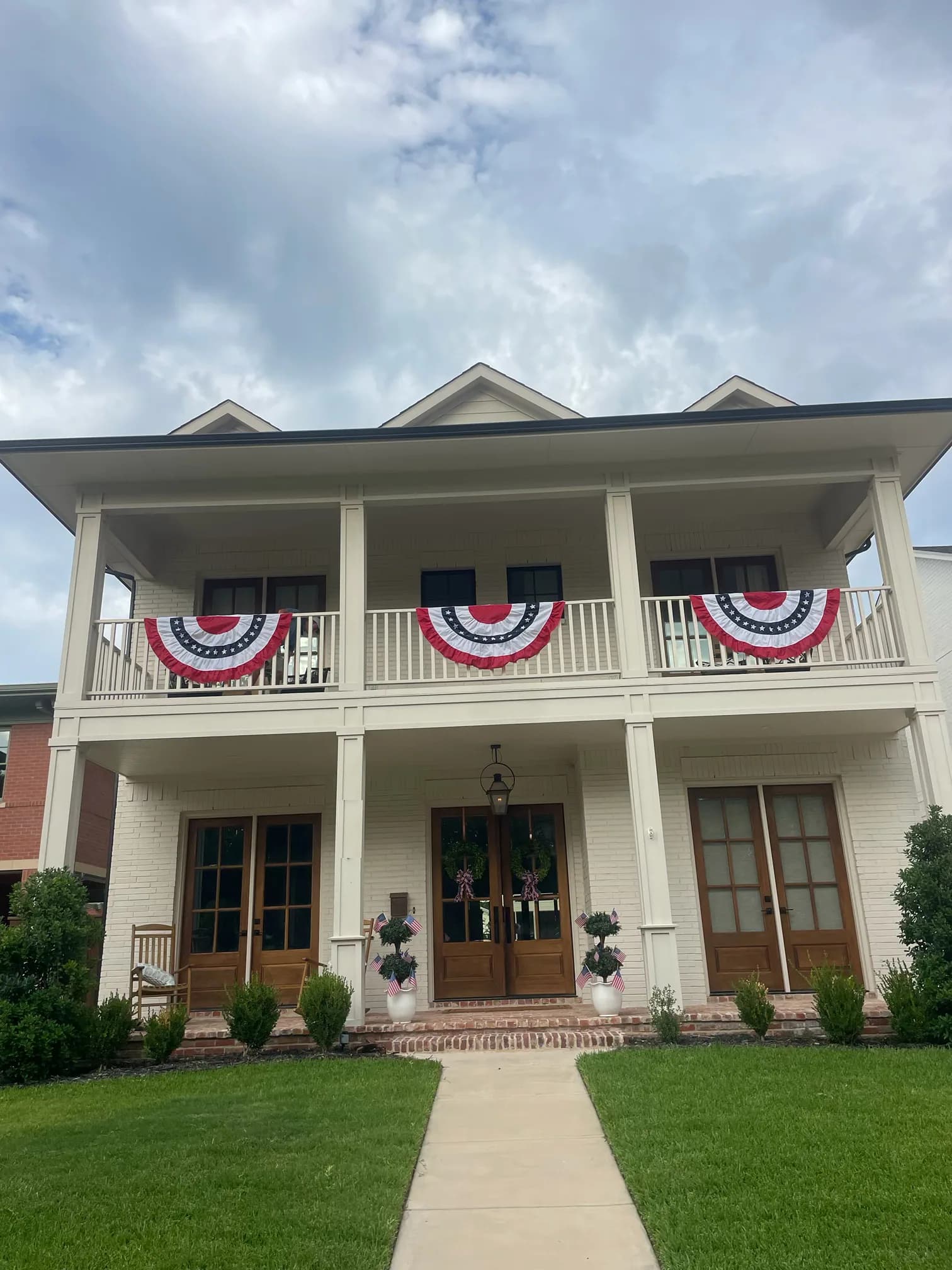 The white brick house with bunting on the upper gallery rail, rocking chairs on the porch and potted topiaries flanking the front doors.