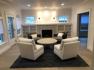 A living room with a tiled fireplace flanked by built-in bookcases, and four pale swivel chairs around a round ottoman on a dark rug.