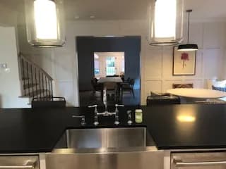 From behind a stainless farmhouse sink with a black counter and bridge faucet, looking through a cased opening to the dining room.