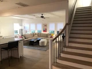 An open living room with a sectional under a row of windows, a ceiling fan, and a stair with a black metal balustrade rising at right.