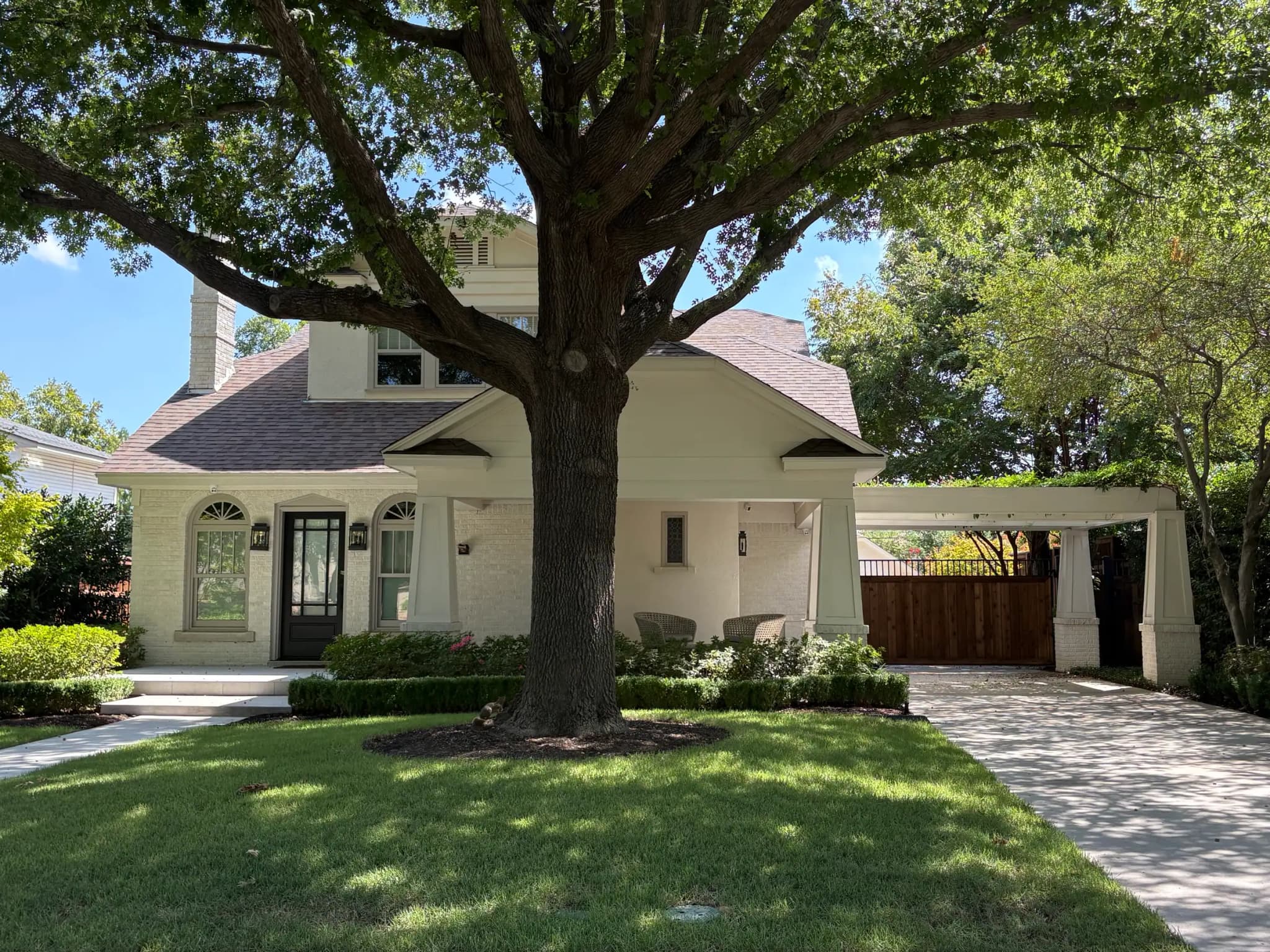 A white-painted brick bungalow under a large spreading tree: a deep front porch on tapered columns, a black front door with arched windows either side, and a matching porte-cochere over the drive.