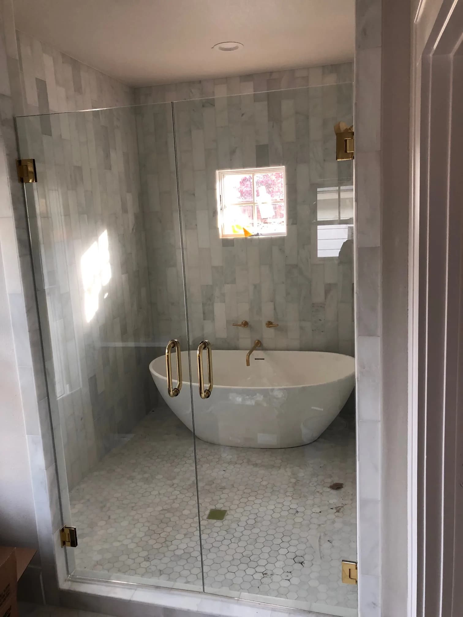 A stone-tiled wet room behind frameless glass with brass hinges and pulls, a freestanding oval tub with a wall-mounted brass filler, and a hexagon mosaic floor still dusty from the work.