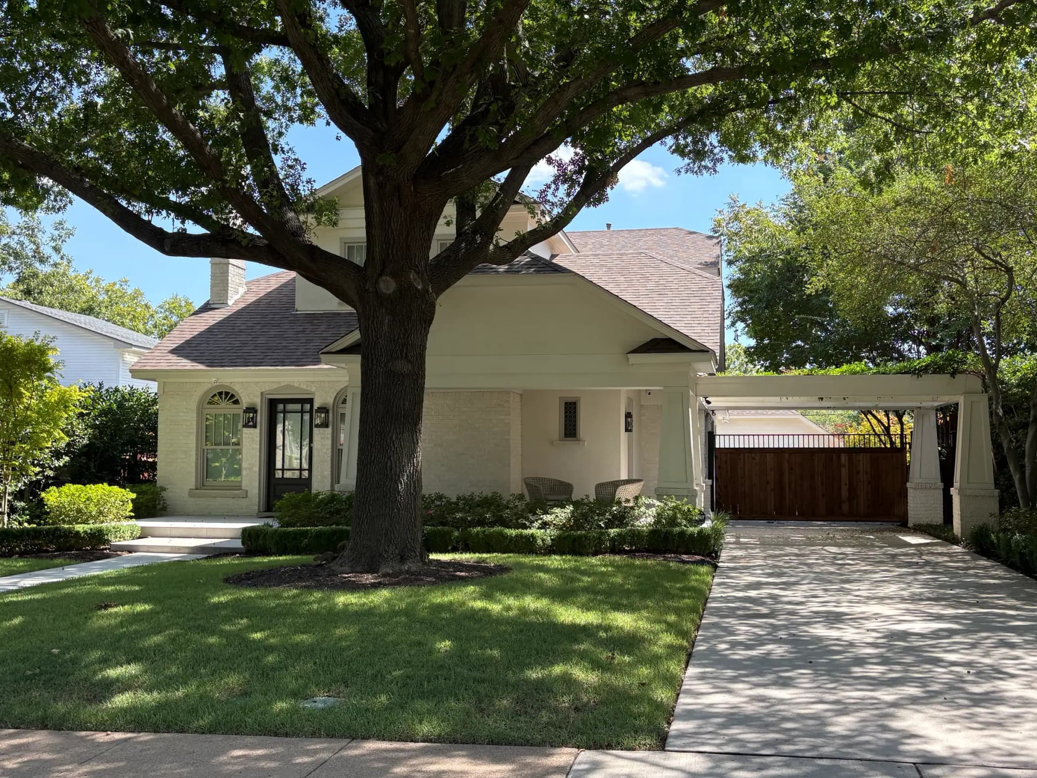 Wider view of the white brick bungalow: the large tree over the lawn, the porch, the porte-cochere and a wood gate to the back.