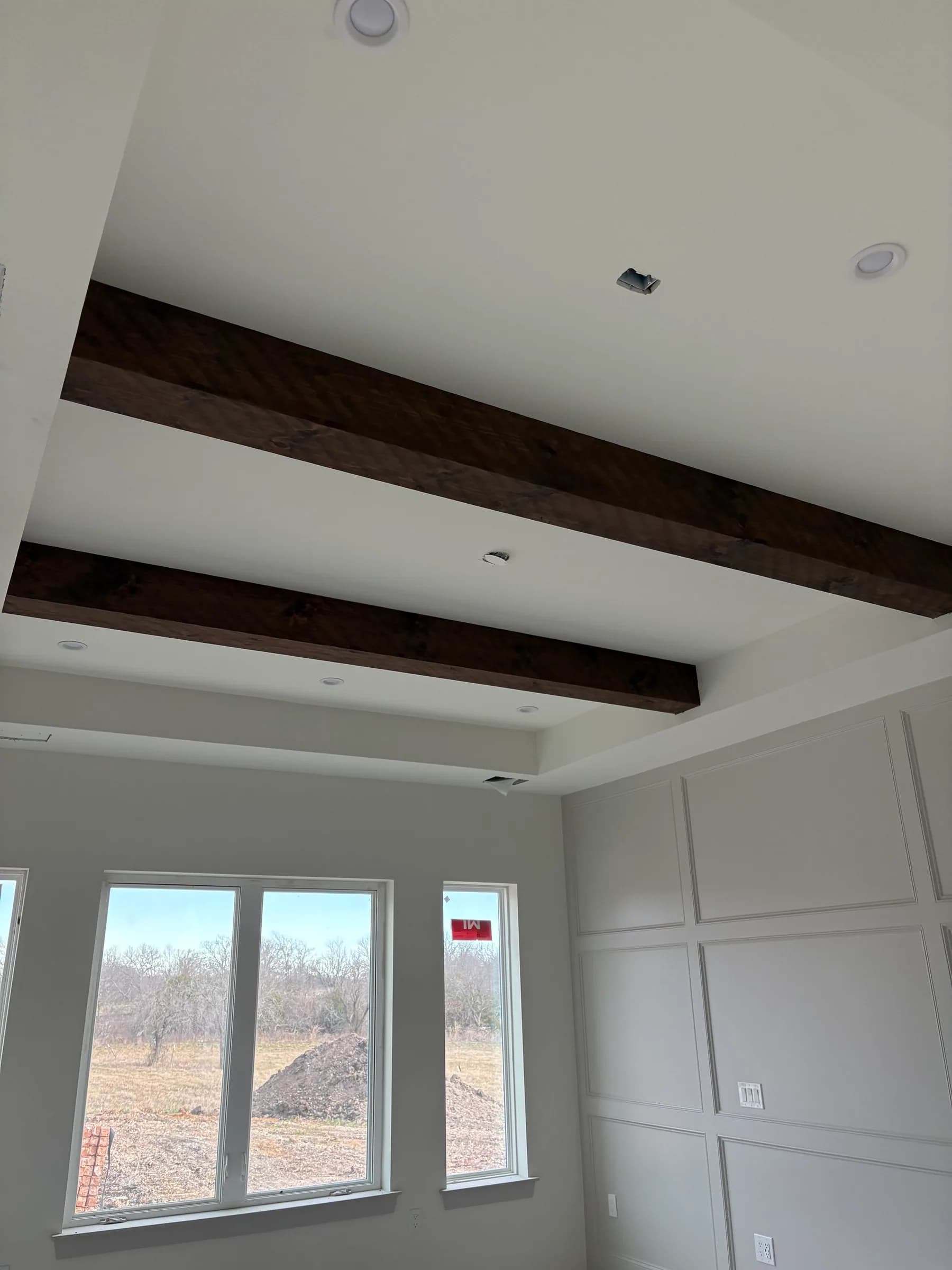 Two stained timber beams across a tray ceiling above a grid-panelled wall, recessed lights roughed in, and windows onto open pasture with a spoil pile outside.