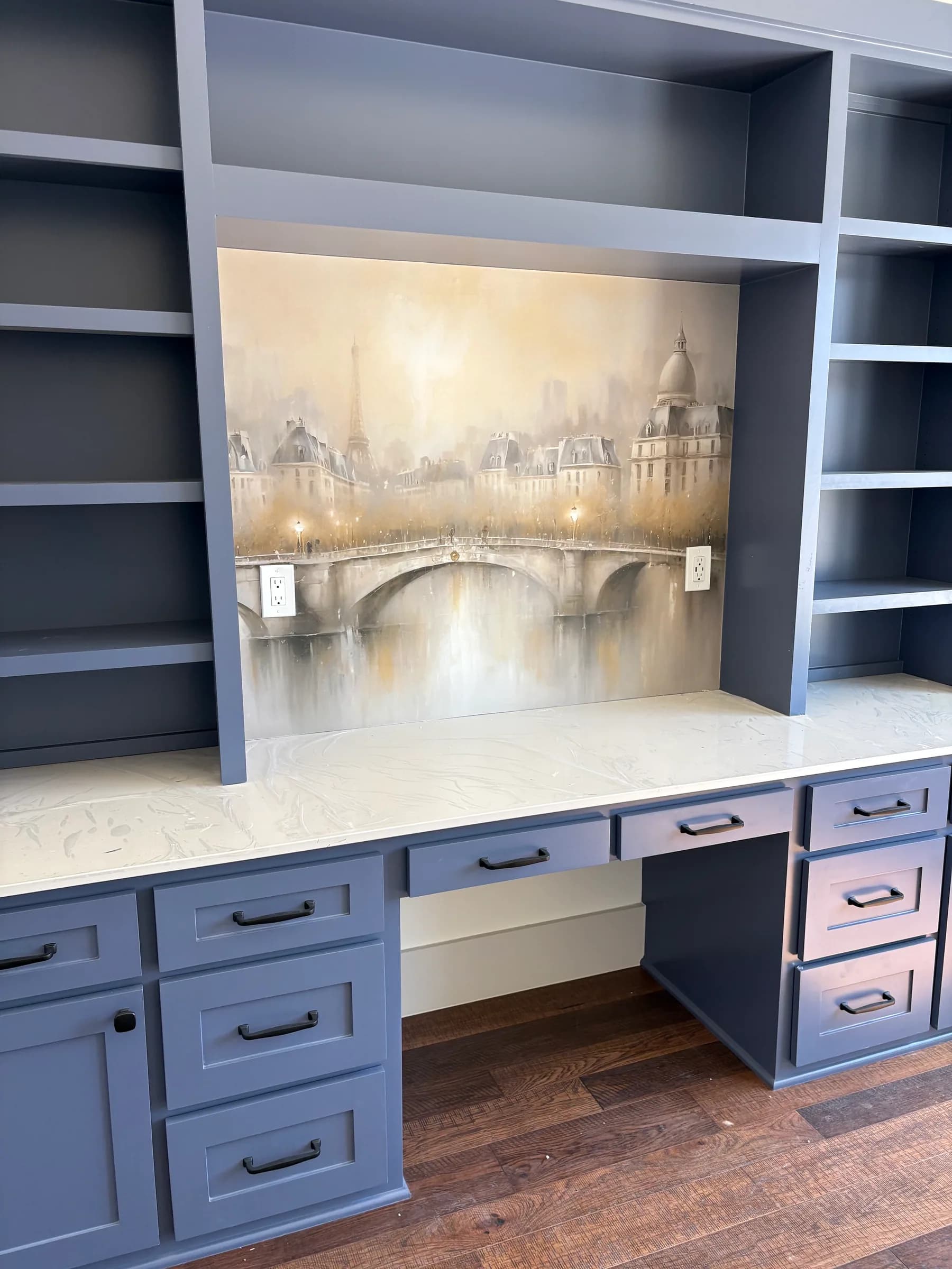 A built-in desk and bookcase wall painted slate blue, with a veined stone top and a canvas of a misty city bridge set into the niche.