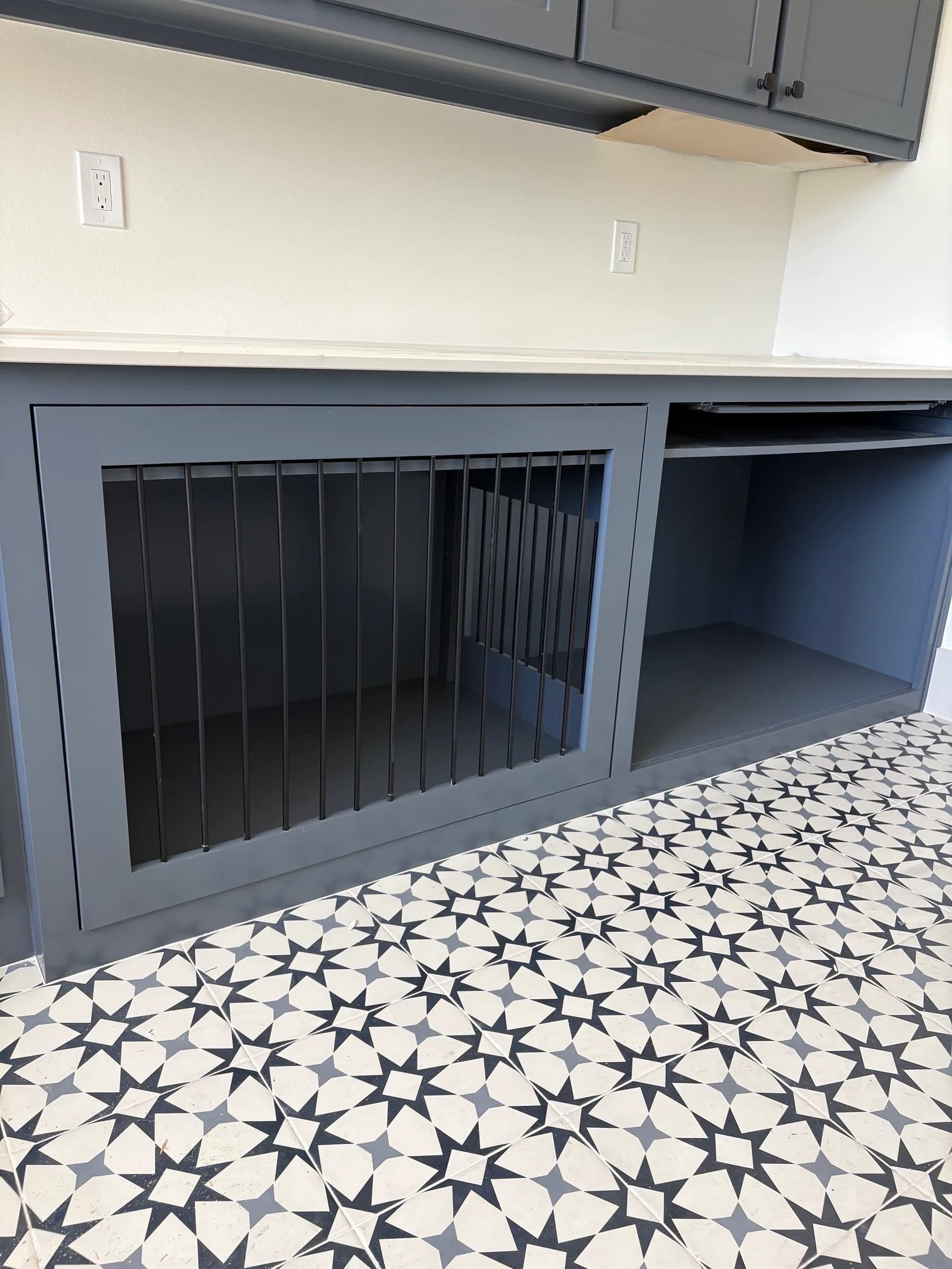 A built-in dog kennel with black metal bars under a laundry counter, slate-gray cabinets above, on a black-and-white patterned tile floor.