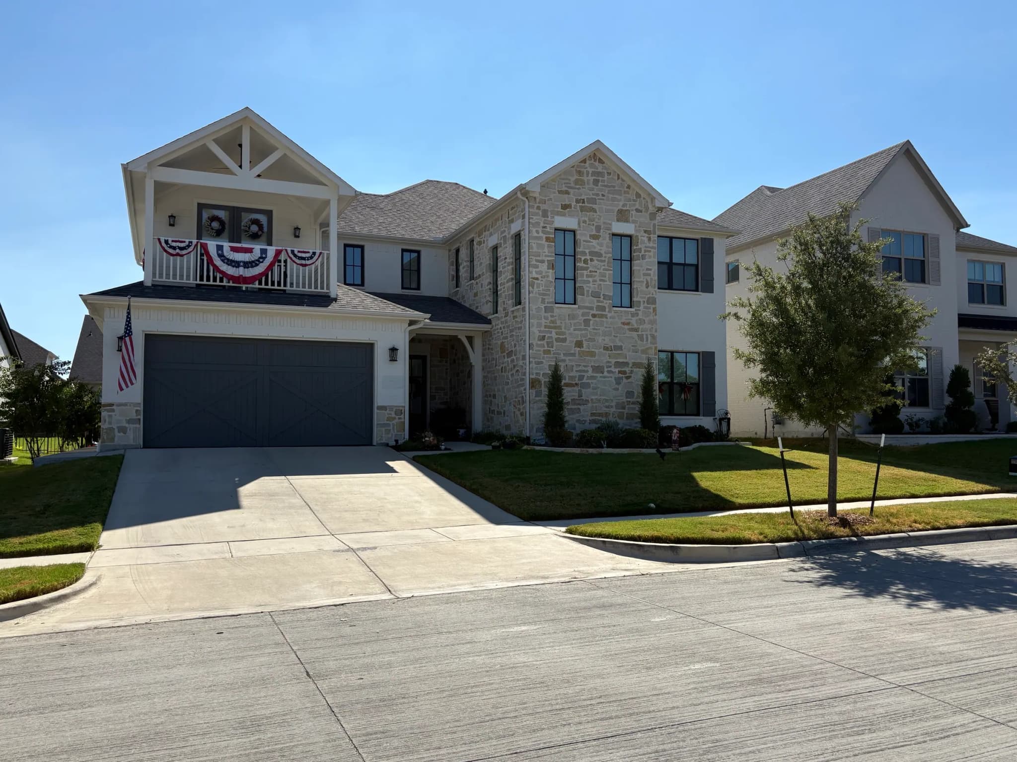 A two-story house of cream stone and white brick with a covered balcony over a gray carriage-style garage door, seen from across the street.