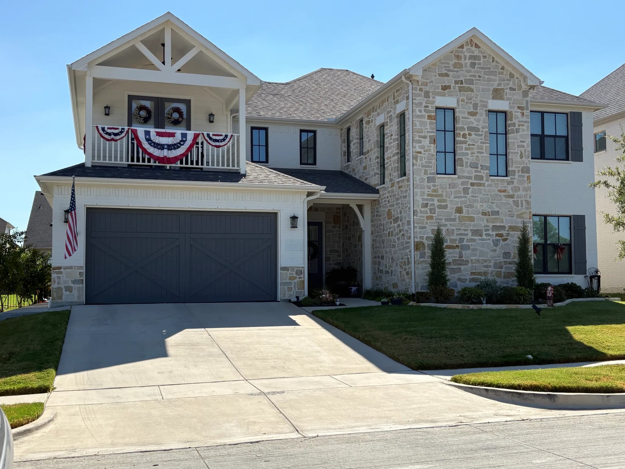 Front elevation: a balcony with a timber truss gable, a gray double garage door, a stone two-story bay and an arched stone entry.