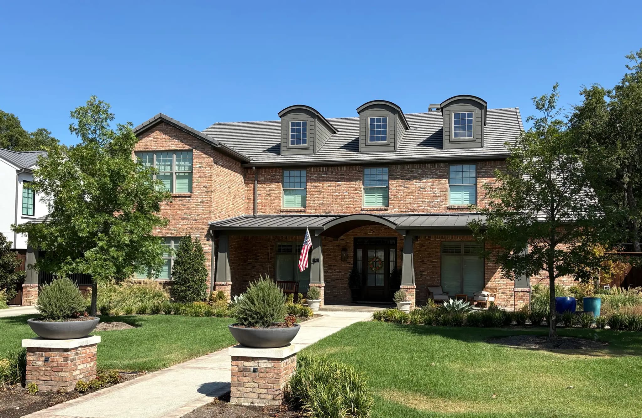 A two-story brick house with three arched dormers, a standing-seam metal porch roof with an arched center, and brick pillar planters at the walk.