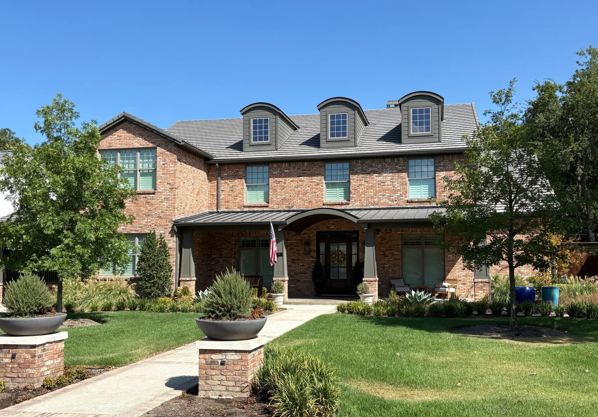 The brick house with arched dormers, a standing-seam porch roof and mature trees either side of the walk.