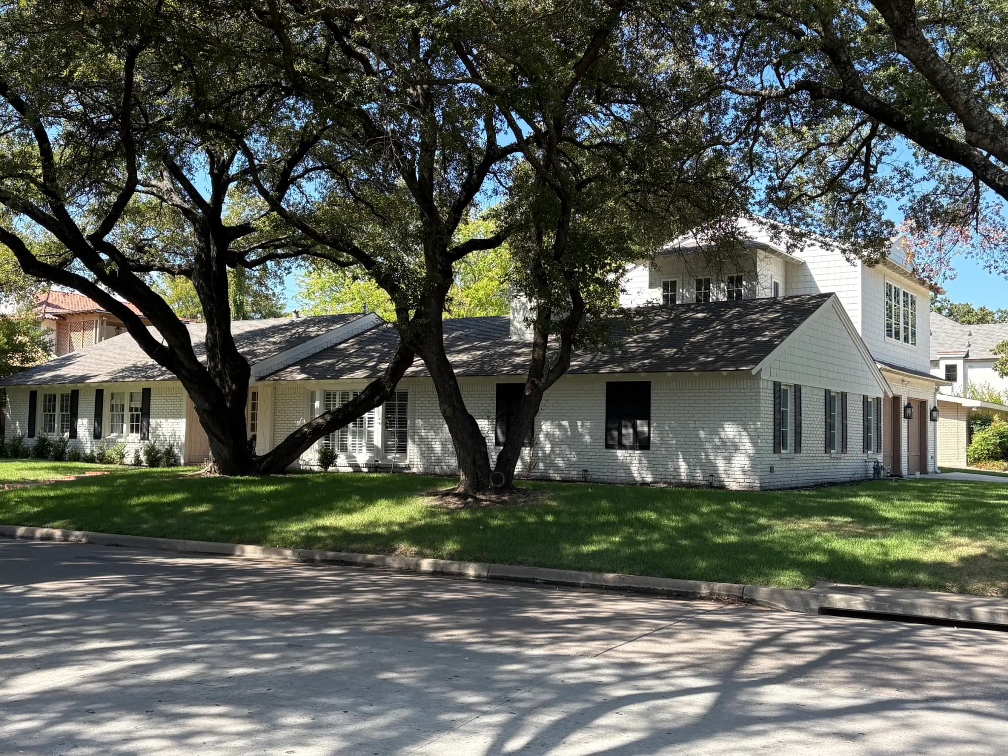 A white-painted brick ranch under big spreading trees, with a shingle-sided second story rising over the garage end.