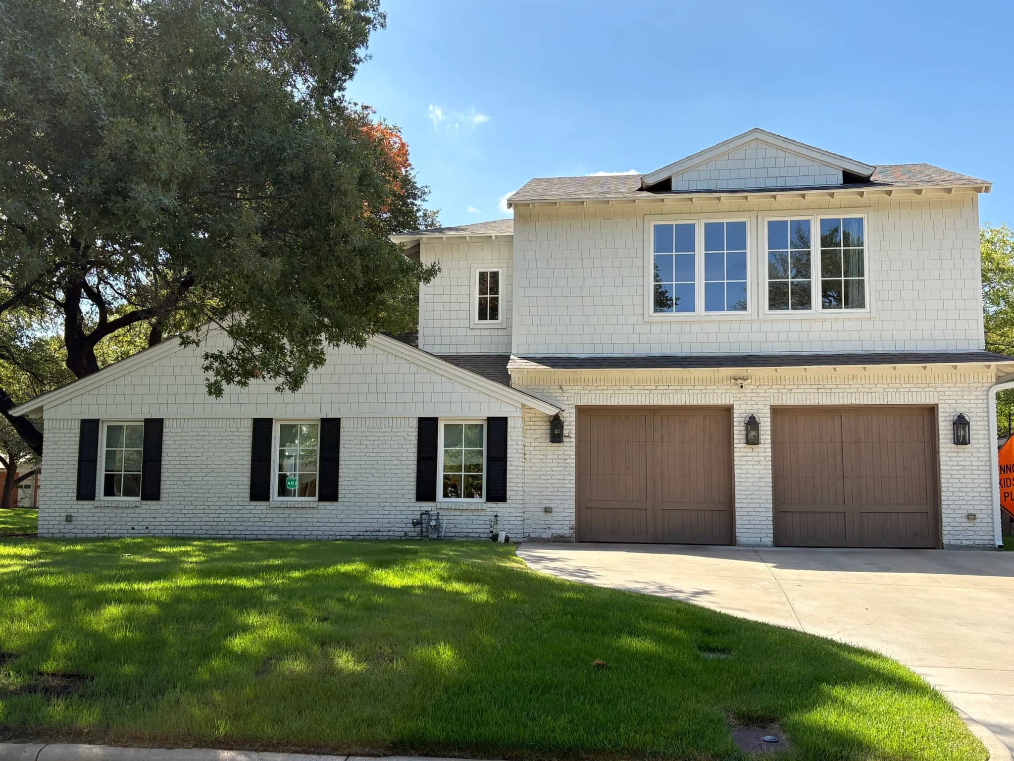 The white brick house from the drive: black shutters, two wood garage doors with lanterns, and the shingle-clad second story above.