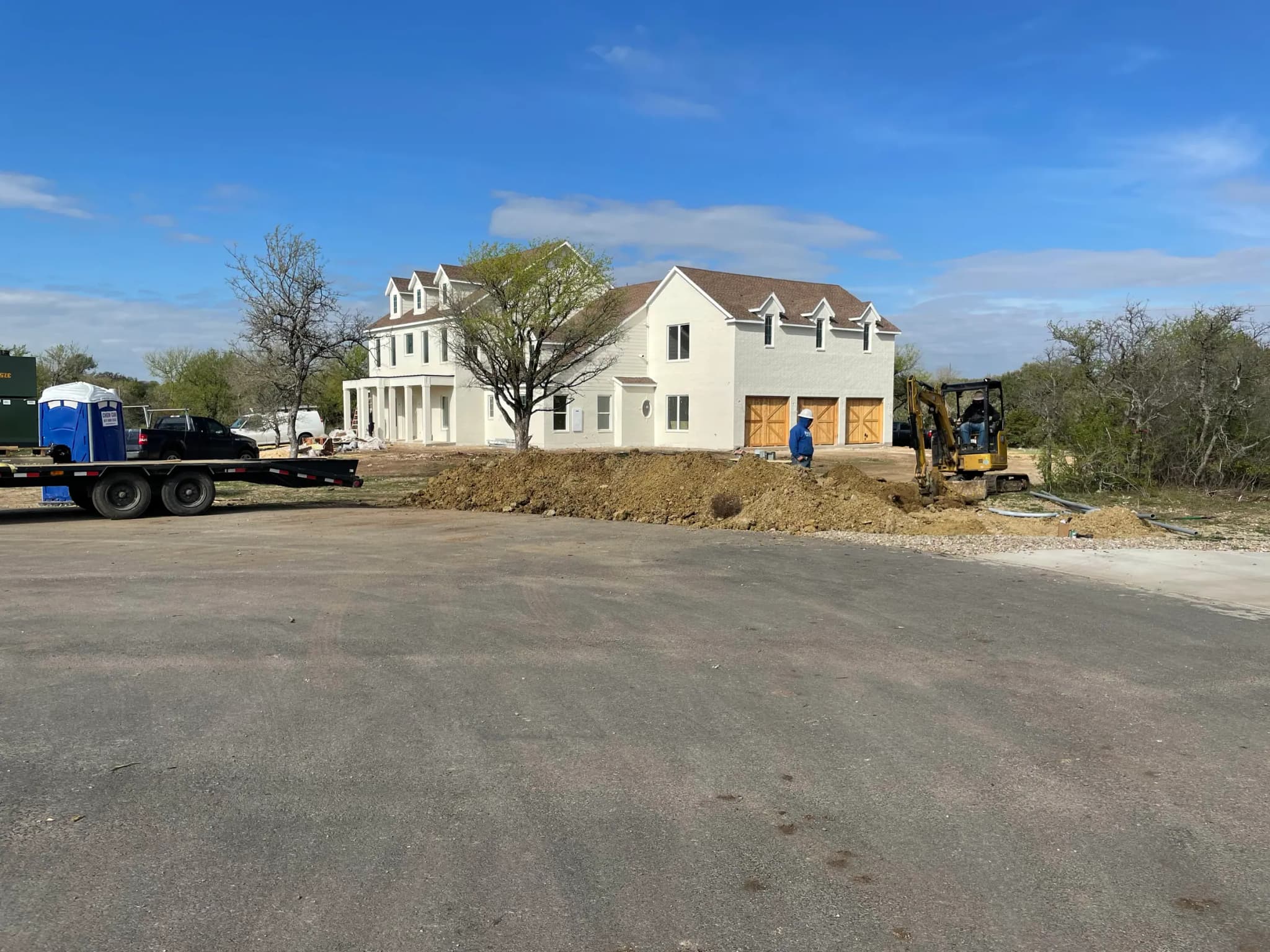 A large white two-story farmhouse with dormers and a three-bay garage, seen across the site during utility trenching: an excavator, a trailer and a spoil pile in front.