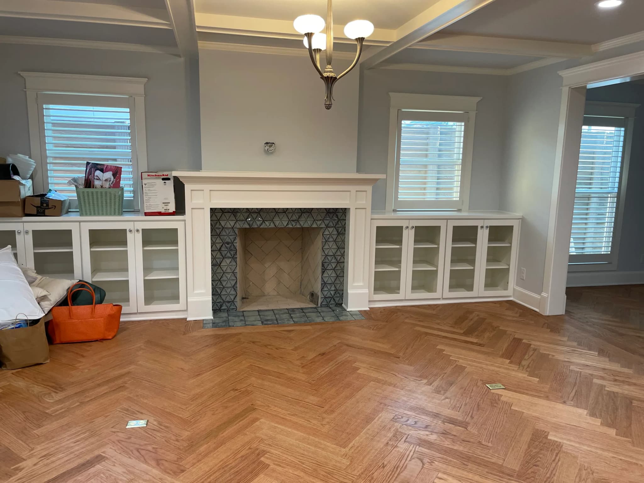 A living room with a coffered ceiling, a patterned-tile fireplace between white glass-front built-ins, and a herringbone wood floor.