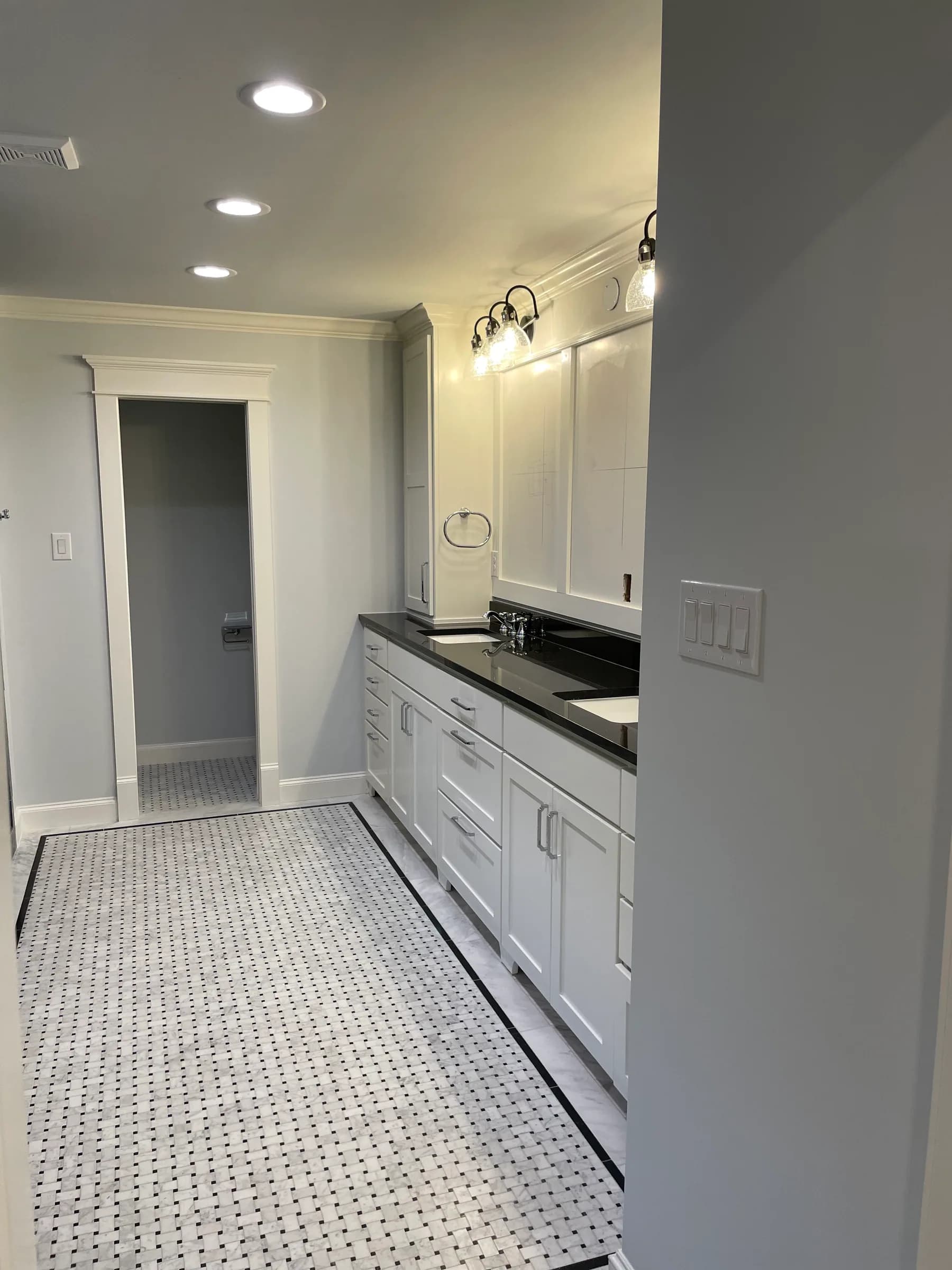 A long bathroom with white shaker cabinets, a dark counter, a basketweave stone floor and a pair of sconces over the mirror.