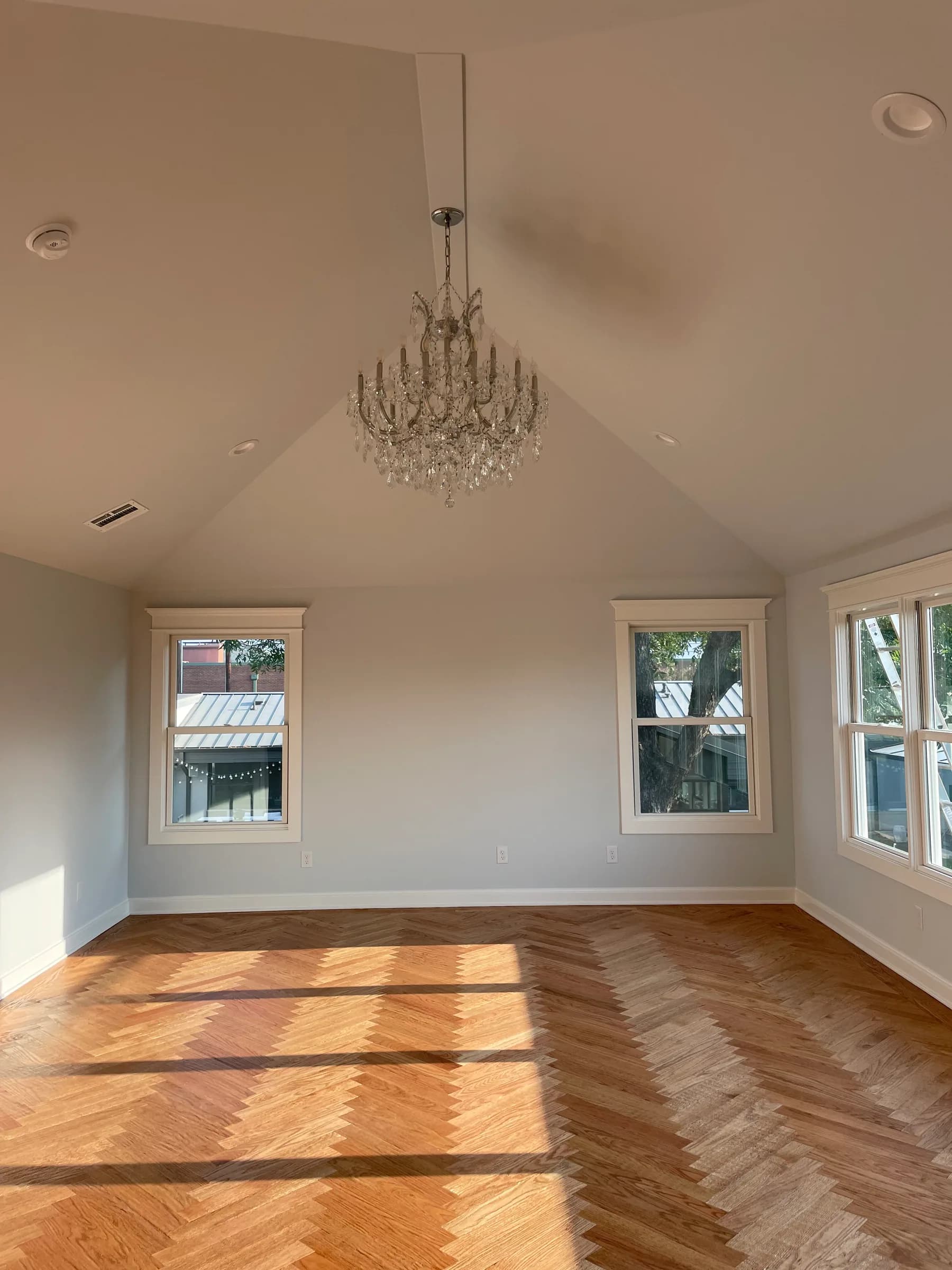 A vaulted room with a glass-drop chandelier, three cased windows and a herringbone wood floor in low sun.