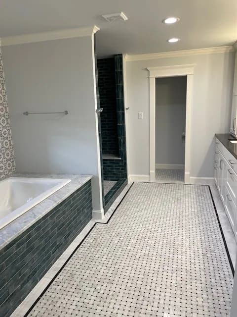 A bathroom with a dark teal subway-tiled tub surround and shower, a stone deck, and a basketweave stone floor with a black border.