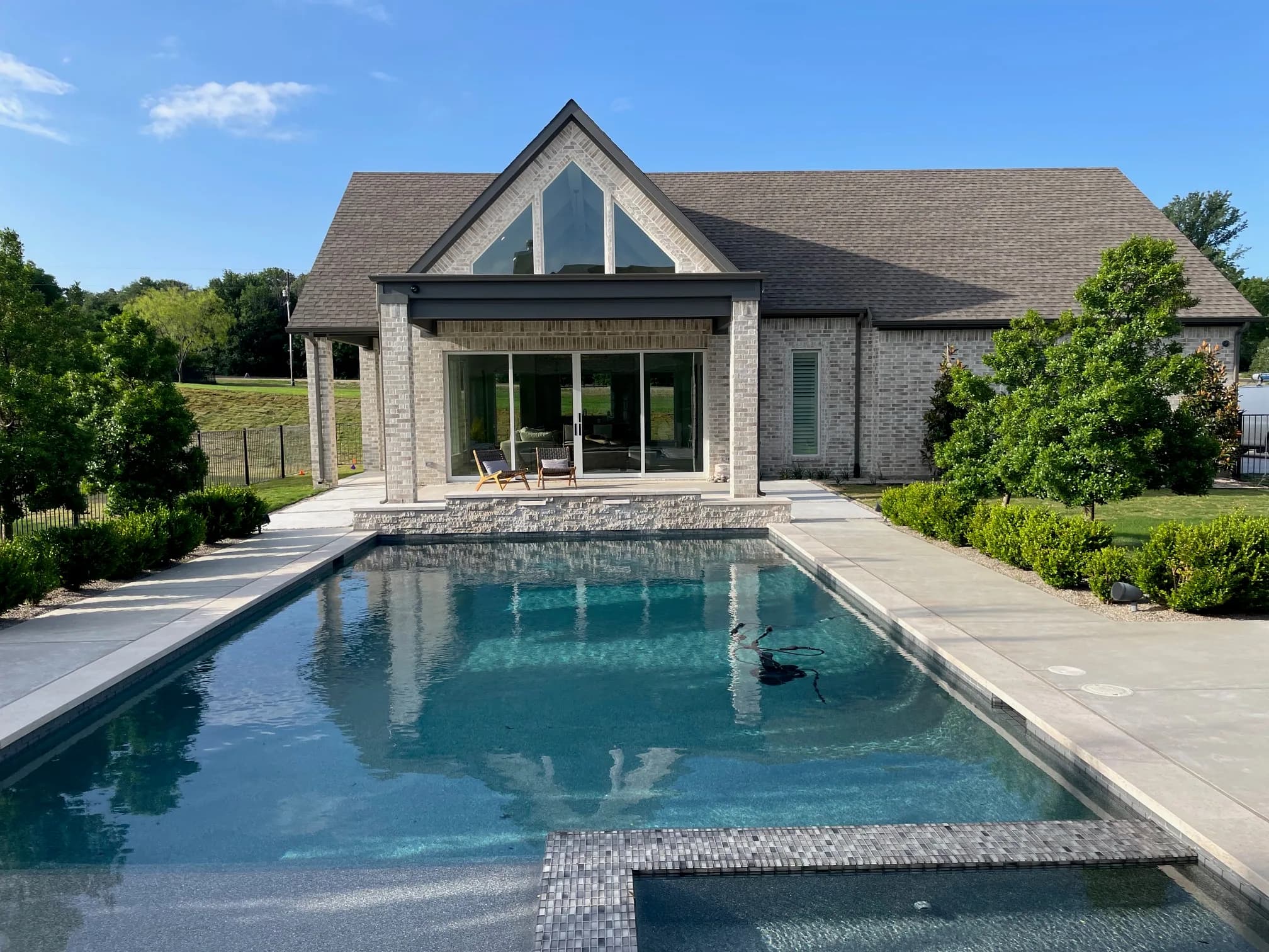 A rectangular pool with a raised spa and a tile-edged tanning ledge, looking back at a blond-brick house with a tall glass gable over a covered patio and sliding doors.