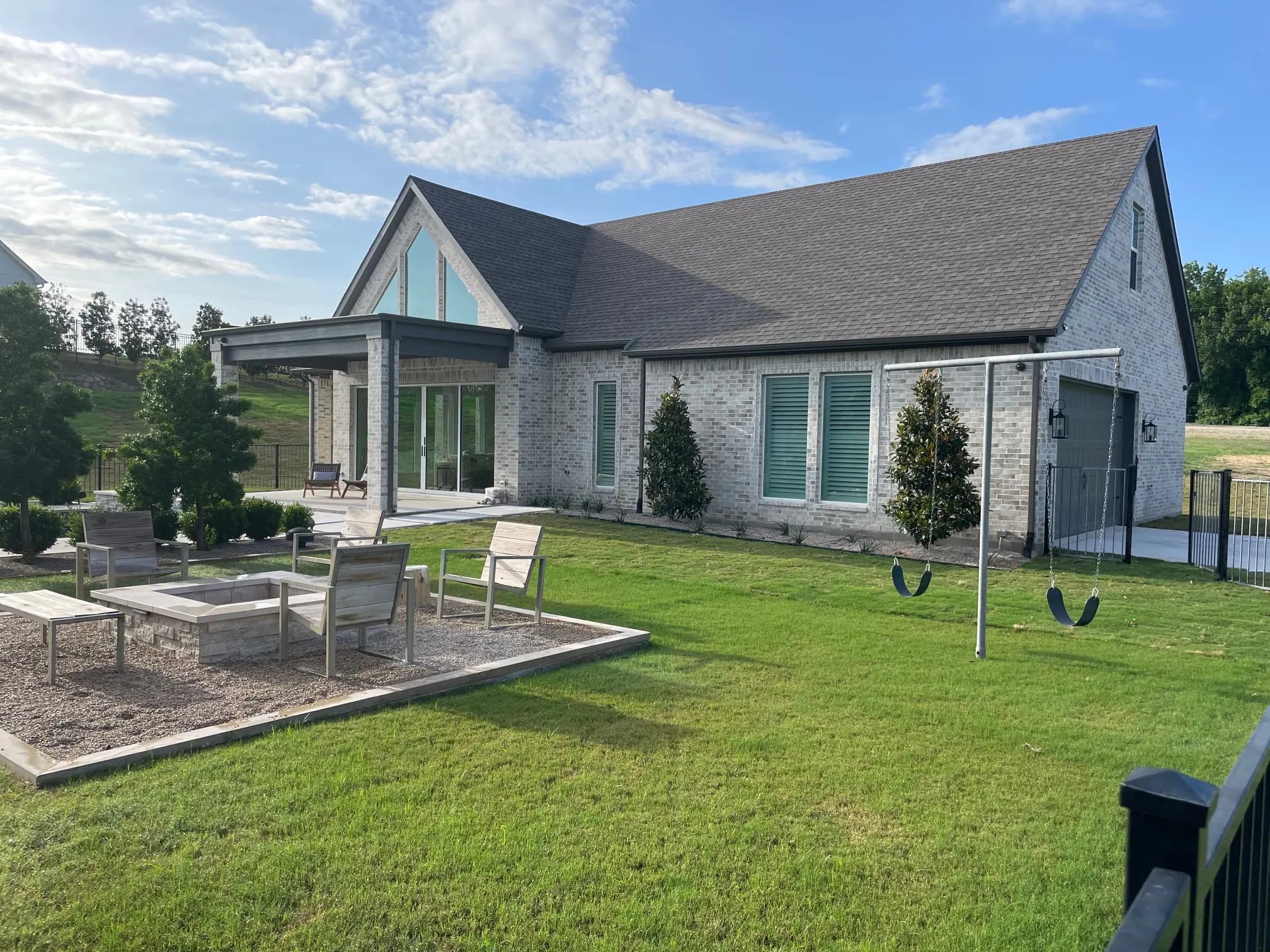 The rear of the house from the lawn: a pale stone fire pit with slatted chairs, a swing set, small trees by the garage and the glass gable above the patio.
