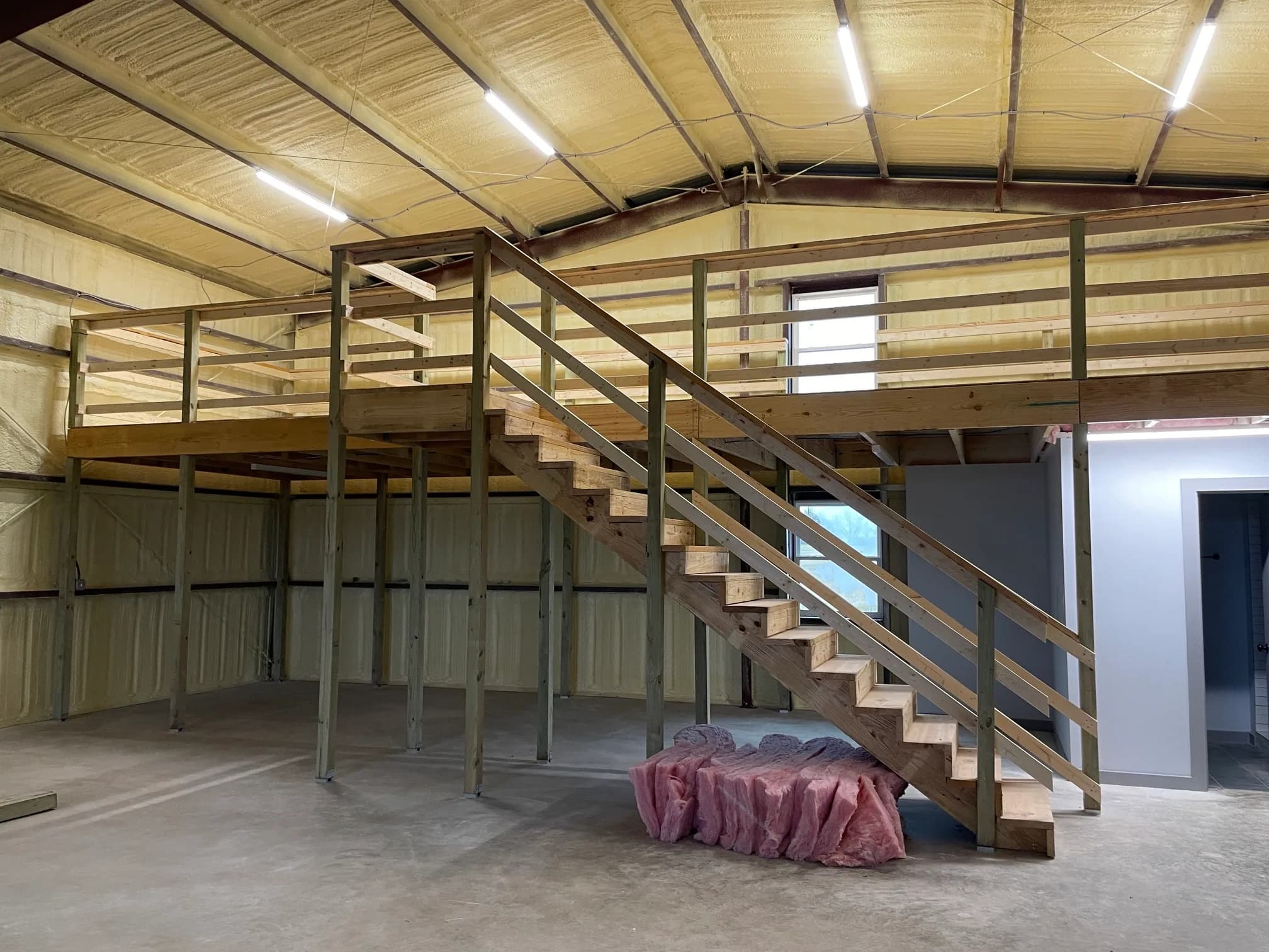 Inside a metal building being finished out: spray-foam insulation on the roof and walls, a timber loft with a stair and rail, and a bundle of batt insulation on the slab.