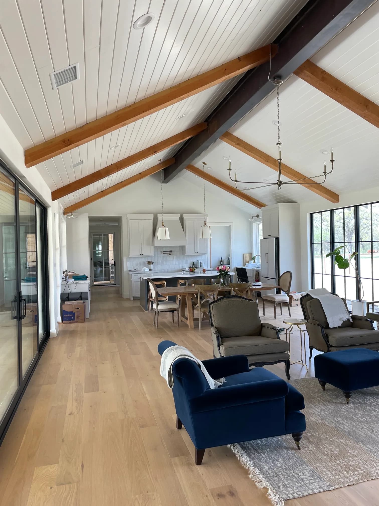 A long vaulted living room under a shiplap ceiling with a steel ridge beam and wood rafters, black steel sliding doors along one wall, pale wood floors, a navy upholstered chair and the kitchen beyond.