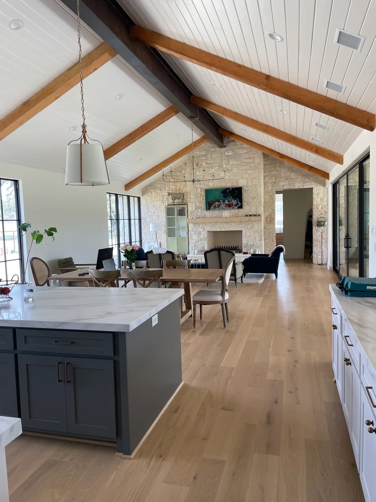 From the kitchen island, dark gray with a stone top under a white drum pendant, looking down the room to a floor-to-ceiling pale stone fireplace wall and black steel windows.