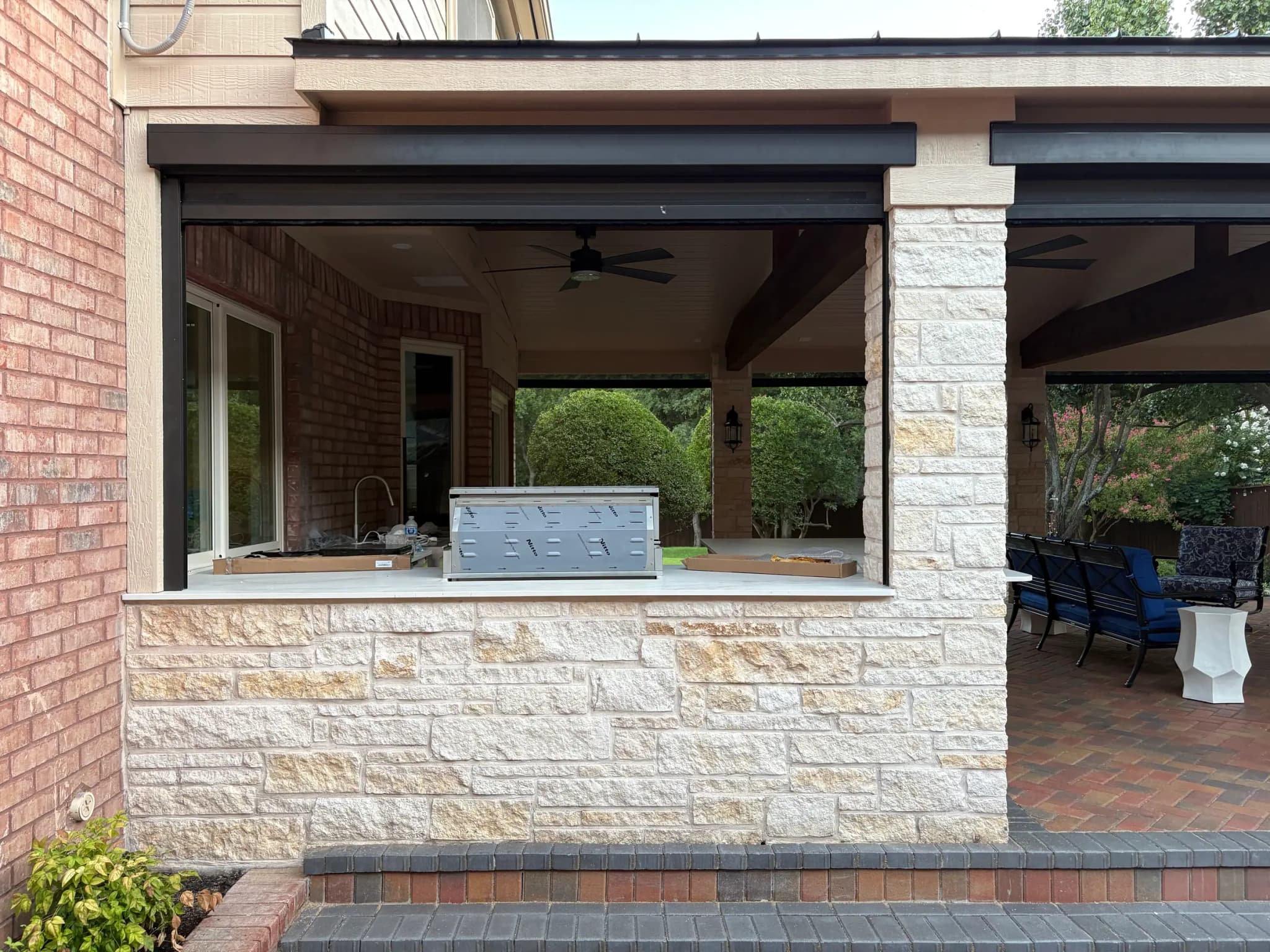 An outdoor kitchen pass-through in pale stone with a stainless grill, under a covered patio with a ceiling fan and retractable screens rolled up.