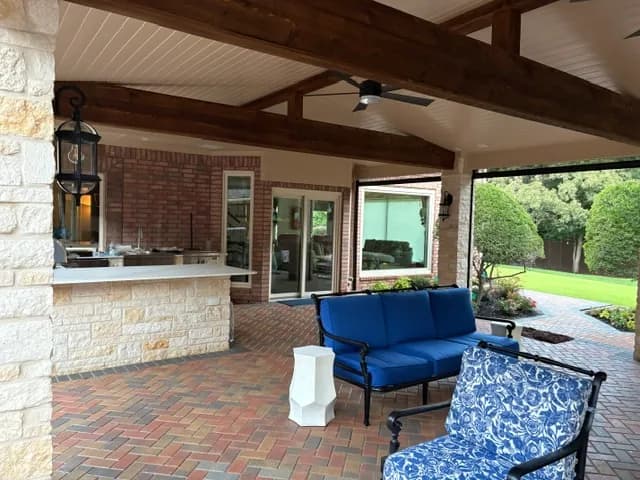 Under the patio roof: timber beams, a plank ceiling, a pale stone bar and a navy cushioned settee on brick pavers, the lawn beyond.