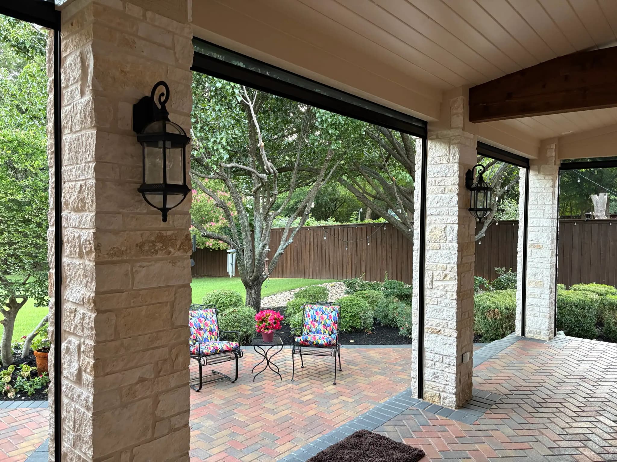 Pale stone columns with black lanterns framing the yard, a tree in pink bloom and a wood fence, and two floral chairs on the pavers.