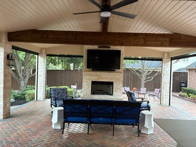 The pavilion's pale stone fireplace with a television above it, timber beams overhead, and a settee and armchairs on herringbone brick pavers.