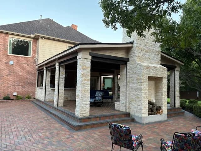 The pavilion from the yard: a pale stone chimney with a wood store, black steel edge beams, and the two-story brick house behind.