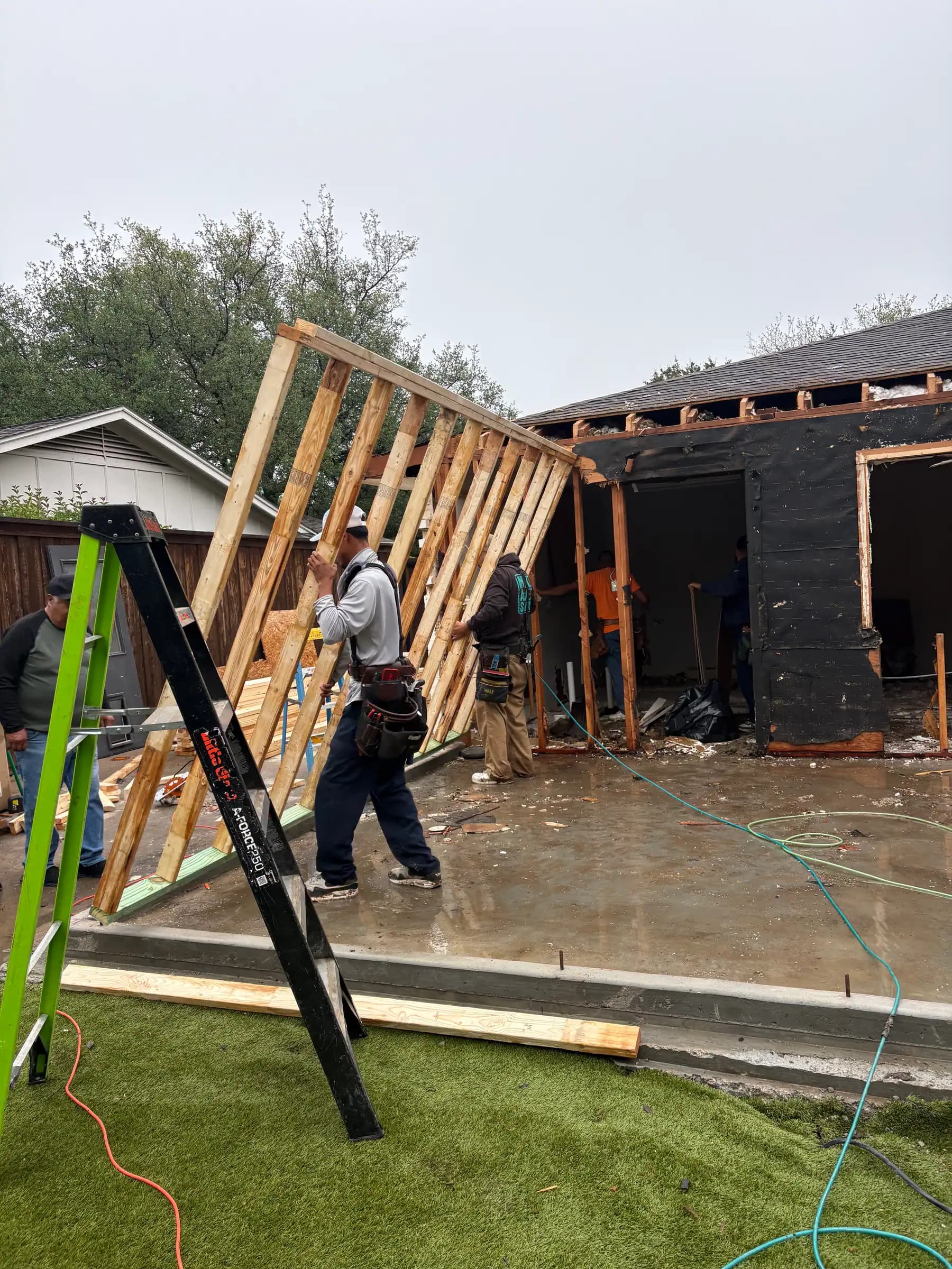 A framing crew standing a stud wall on a new slab beside an existing house opened up for the addition, the ground wet around the slab.