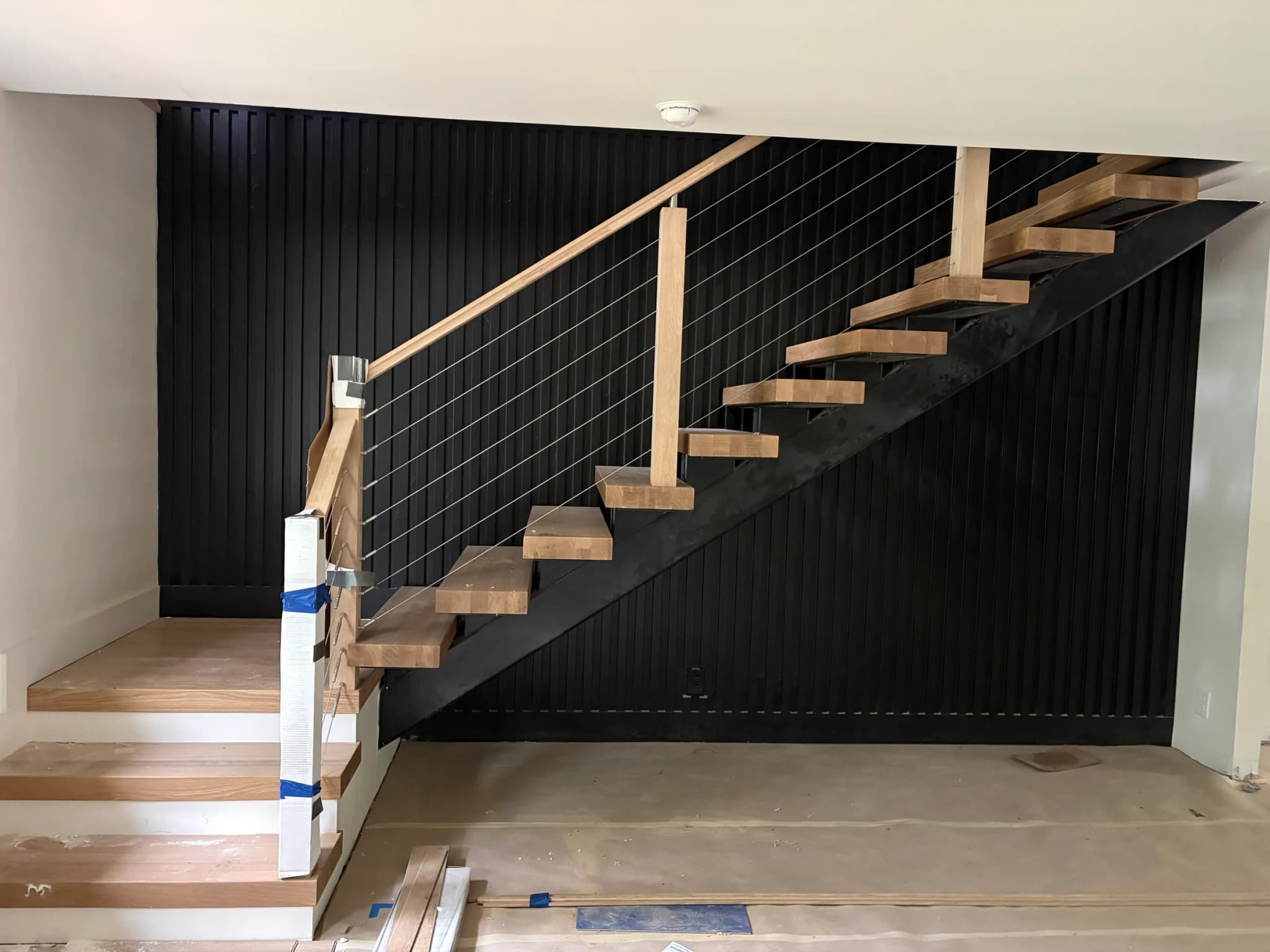 A floating pale wood stair on a black steel stringer with cable rails, against a wall of black vertical slats, the treads still taped for protection.
