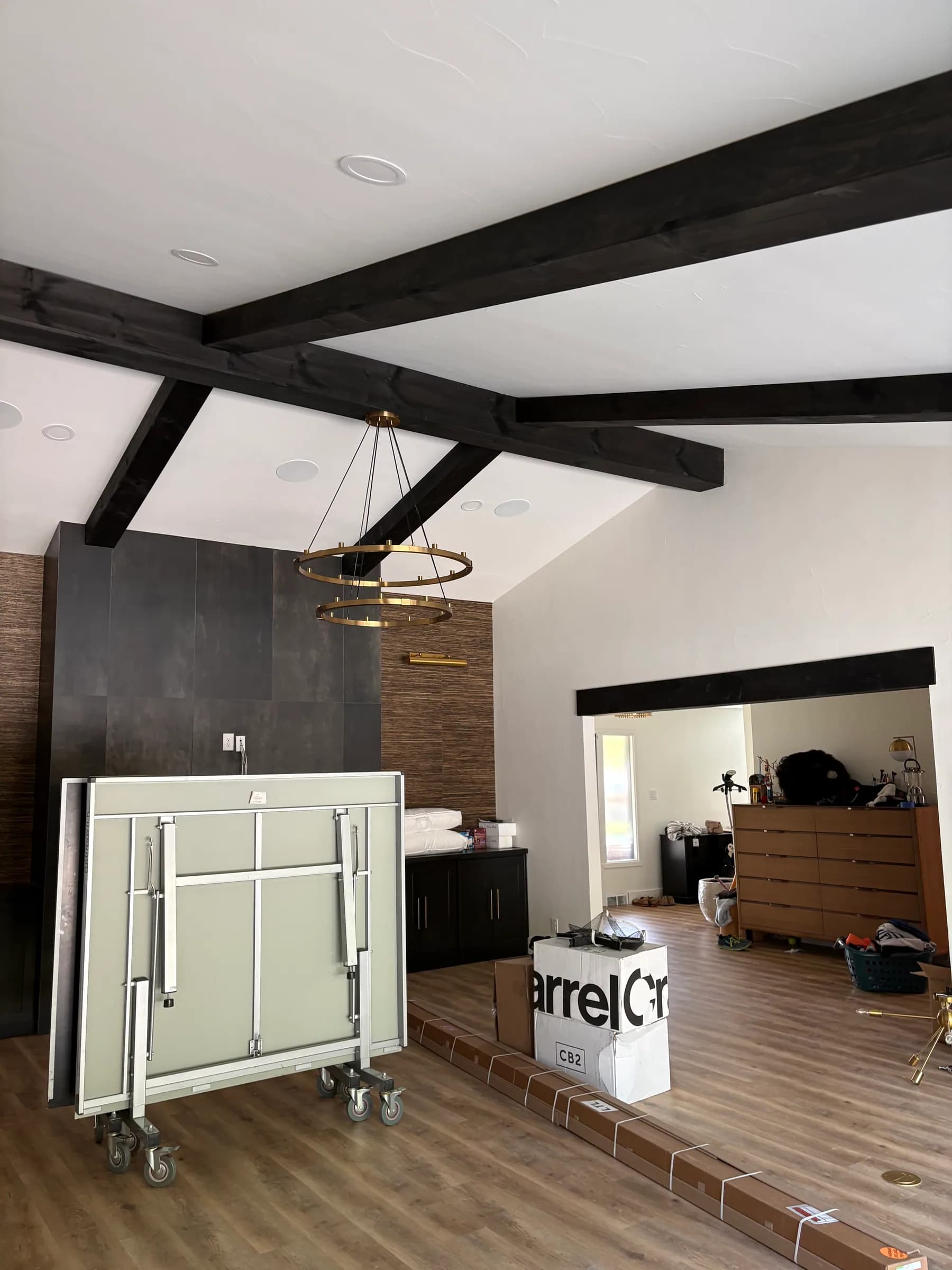 A vaulted room with dark-stained beams, a brass ring chandelier and a dark large-format tile feature wall, mid move-in.