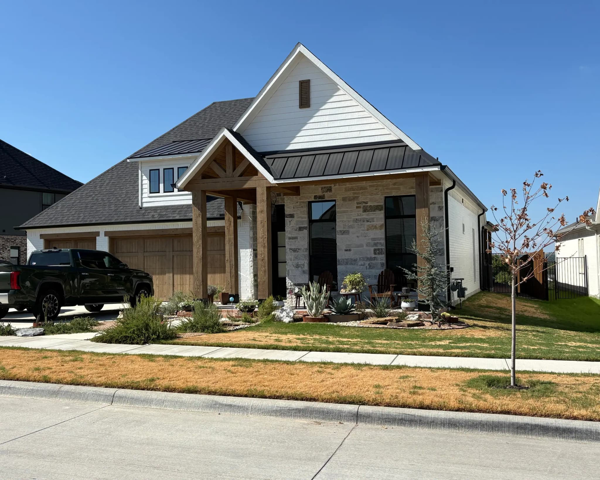 A modern farmhouse with a timber entry gable, a standing-seam metal accent roof, stone at the entry and a two-bay wood garage door, seen from the street.