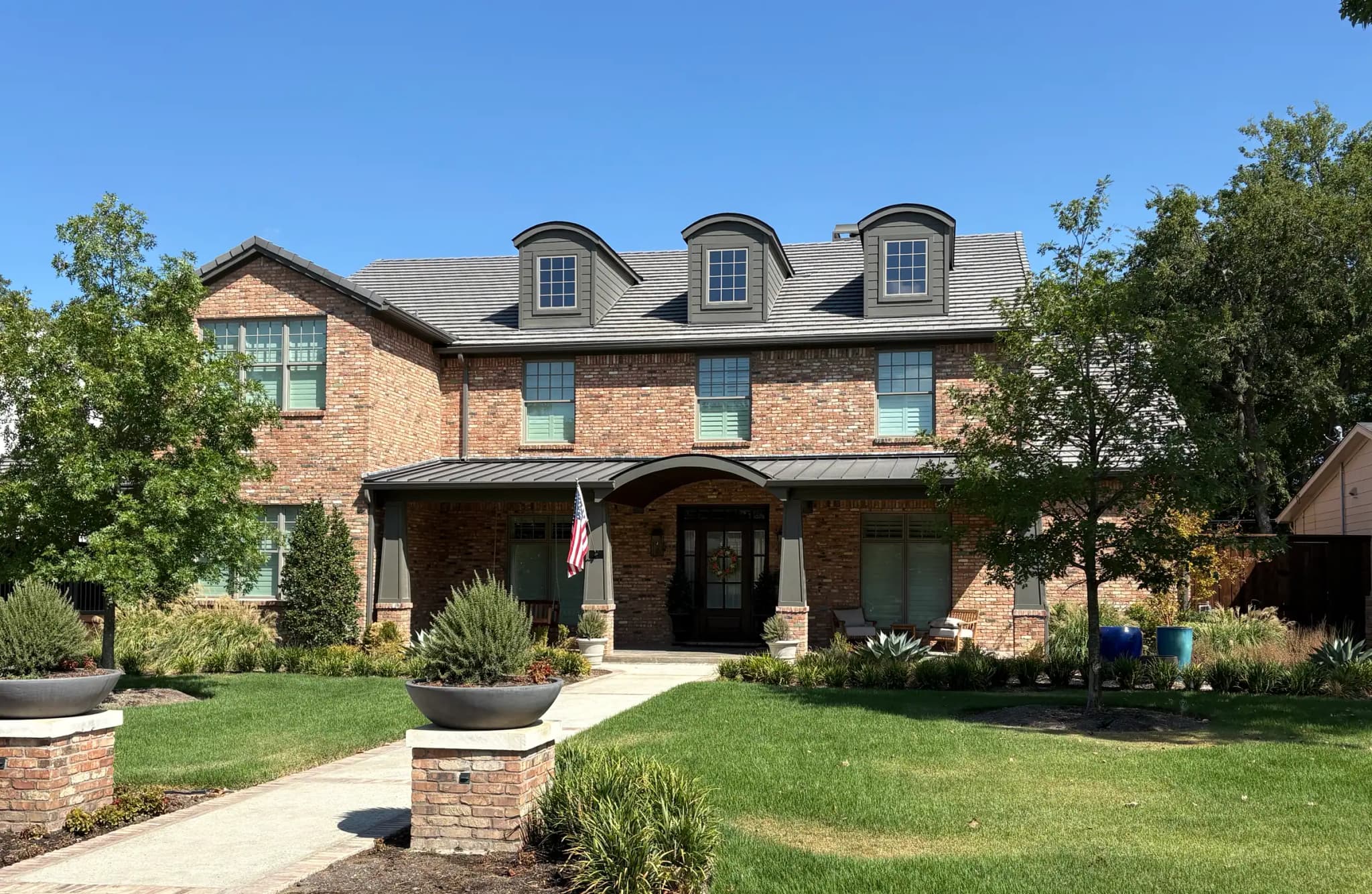 A two-story brick house with three arched dormers, a standing-seam metal porch roof with an arched center, and brick pillar planters at the walk.