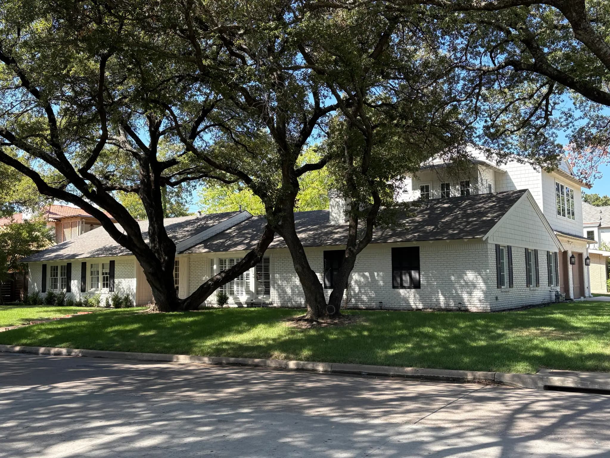 A white-painted brick ranch under big spreading trees, with a shingle-sided second story rising over the garage end and a wide drive curving to it.