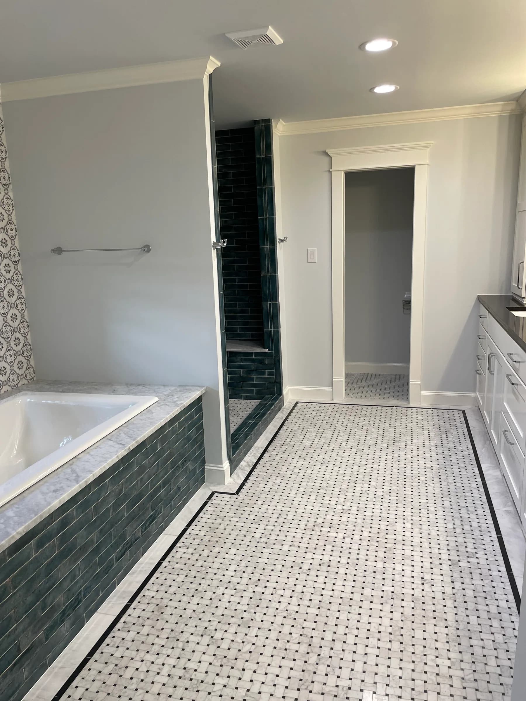 A long bathroom with a drop-in tub under a stone deck, a dark teal subway-tiled shower behind a glass panel, and a white mosaic floor with a black border.