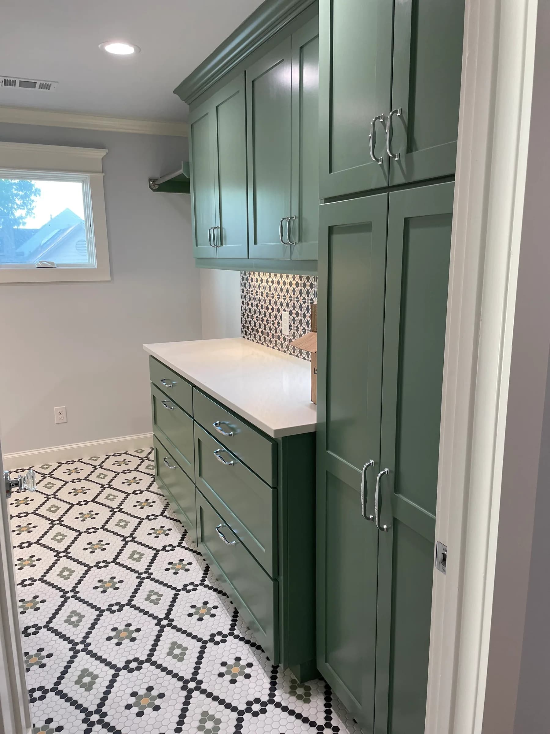 A laundry room in deep green cabinetry with a pale stone counter, an open shelf on black brackets and a patterned tile floor.