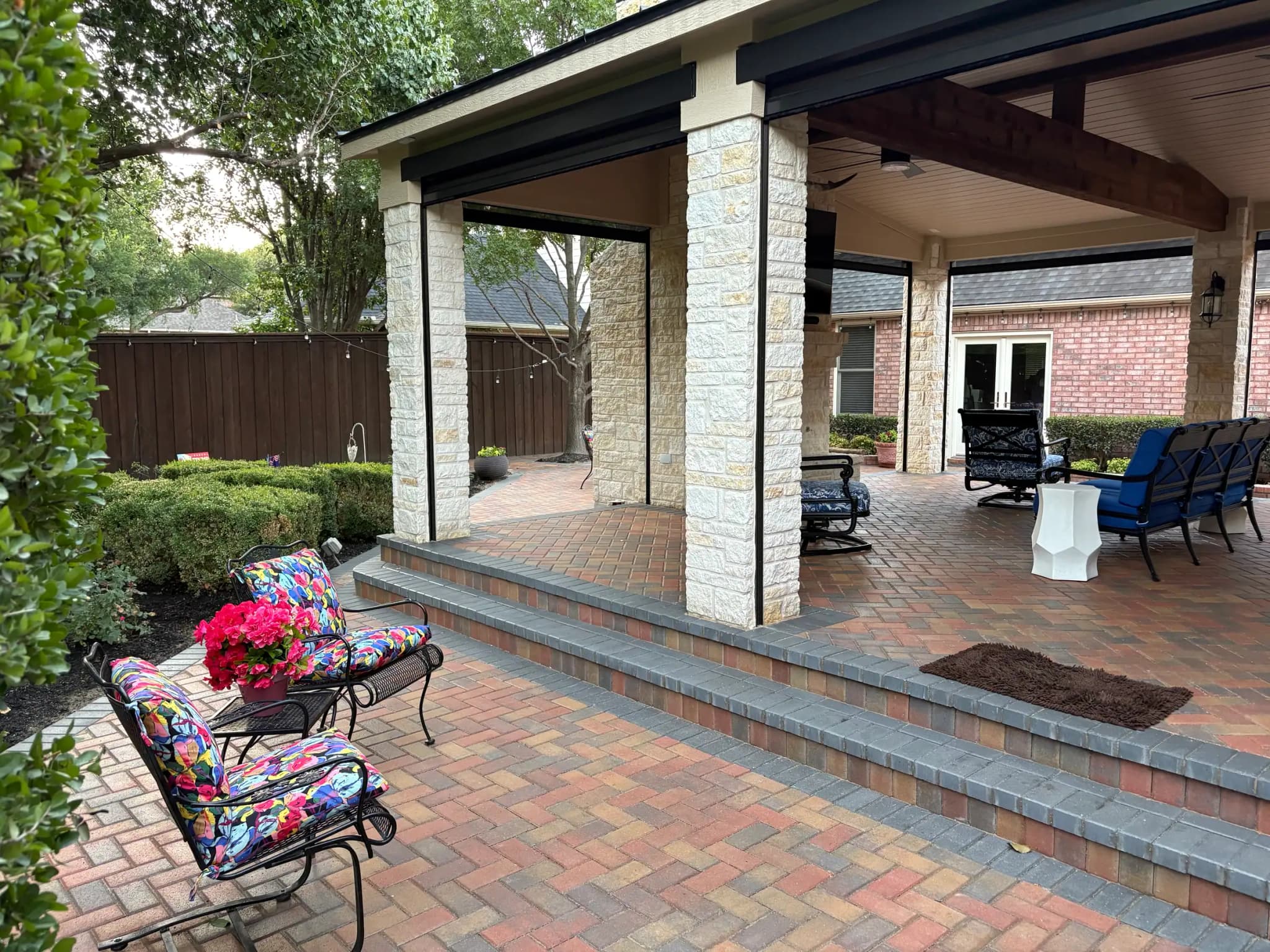 The covered pavilion from the garden: pale stone columns on a raised paver floor, steps up to the seating, and clipped hedges along the bed.