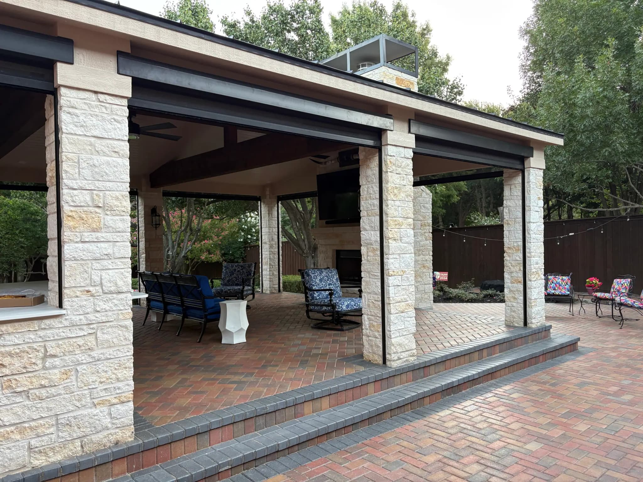 The pavilion from the side: pale stone columns under dark beams, a raised herringbone paver floor, and seating along its length.