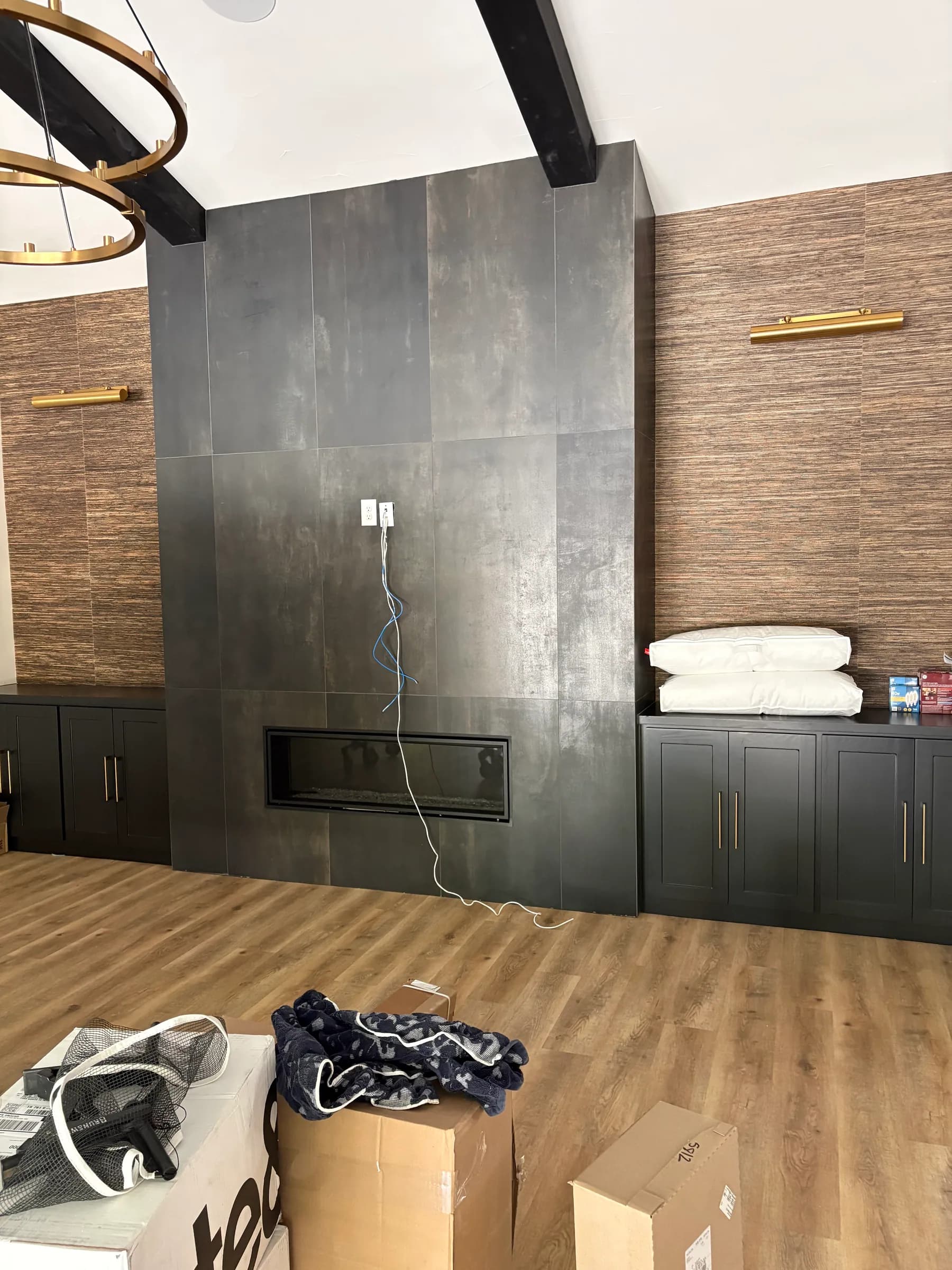 A vaulted room with a dark large-format tile fireplace wall, a woven-textured wallpaper panel with a brass picture light, black cabinets below, and wood floors just laid.