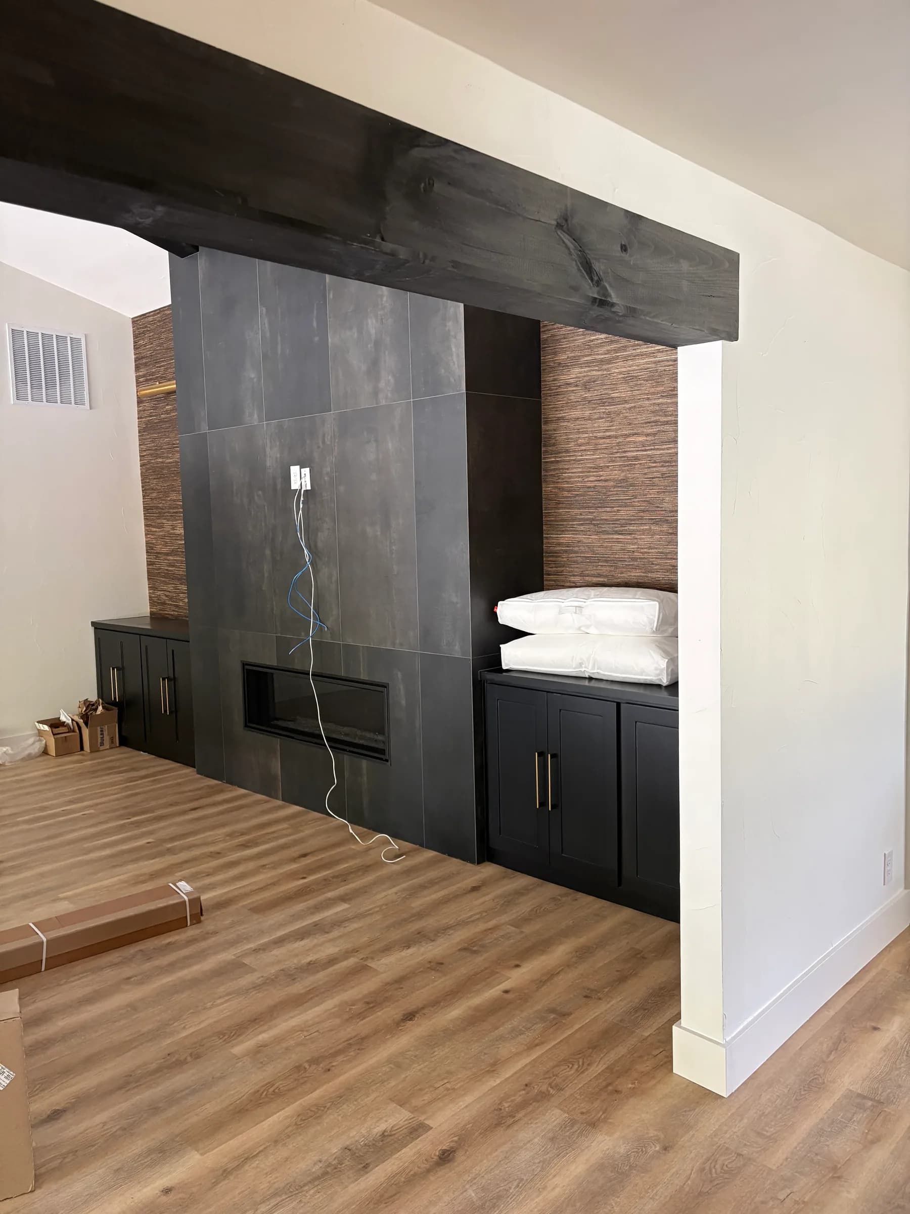 The same room from further back: a stained timber beam across the ceiling, the tiled fireplace wall between black cabinets, and a bench seat under the woven-textured wallpaper panel.