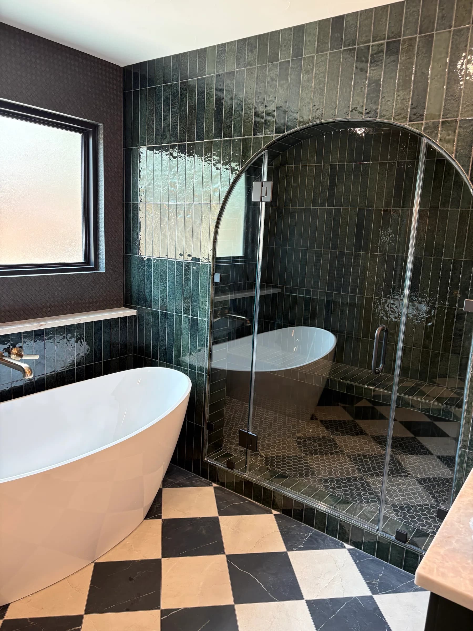 A bathroom in dark glazed stacked tile with an arched glass shower door, a freestanding tub and a checkerboard stone floor.