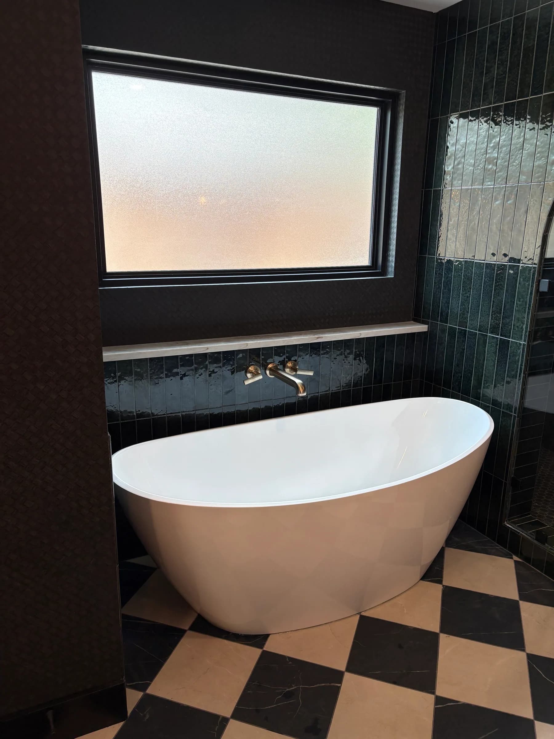 The freestanding tub below a frosted window with a wall-mounted filler, against dark glazed tile and a checkerboard stone floor.
